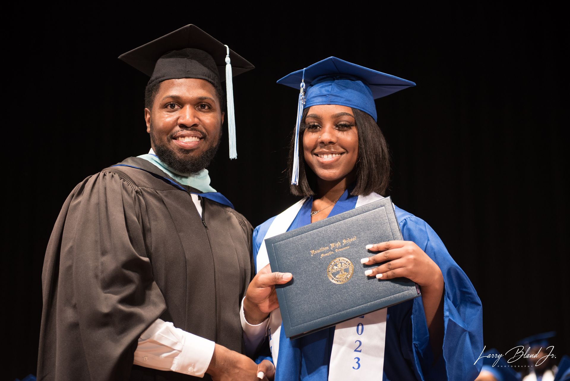 Two people at a graduation ceremony: one in black regalia, the other in blue, holding a diploma, smiling.