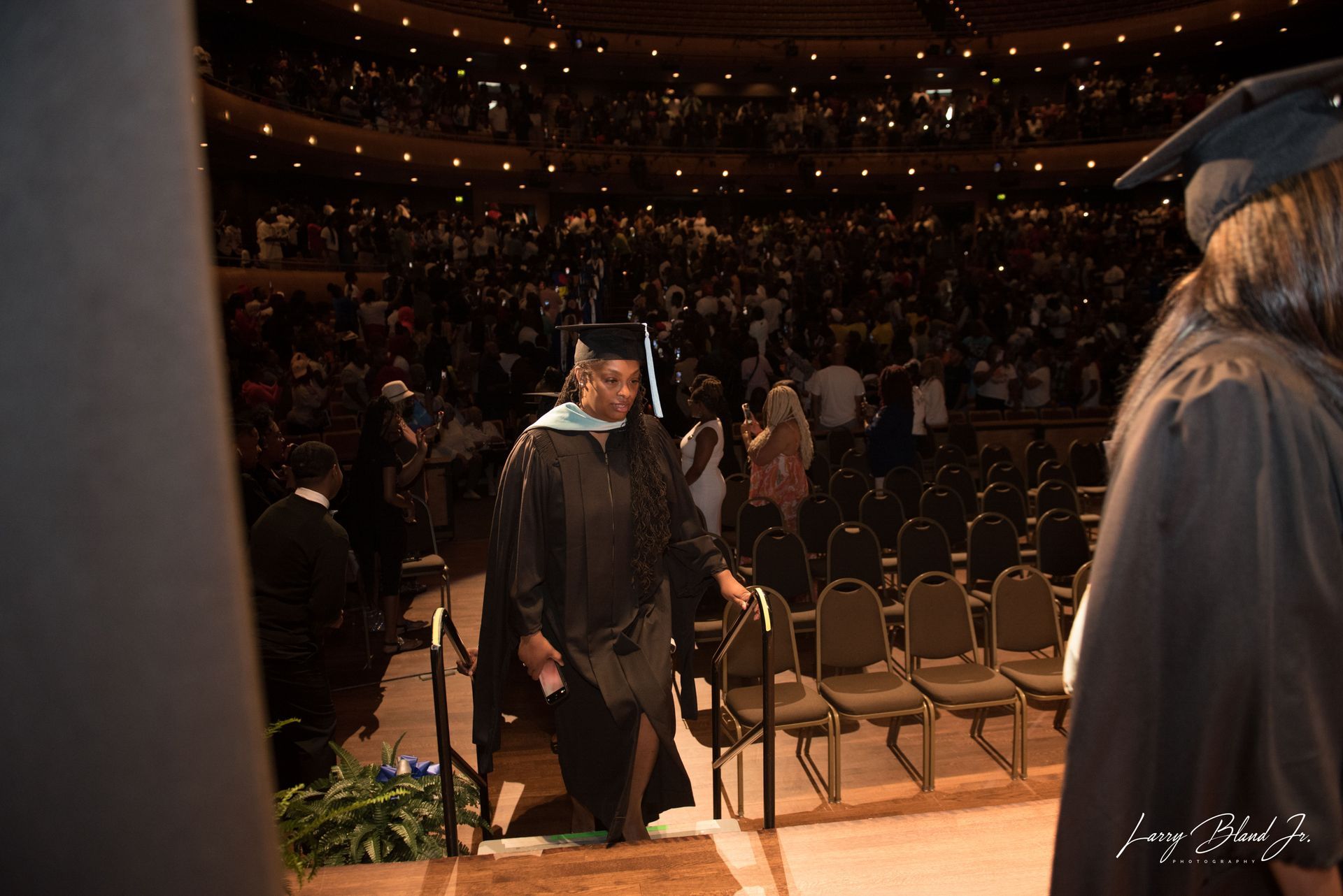 Graduate descending steps in a graduation ceremony, with a crowded auditorium in the background.