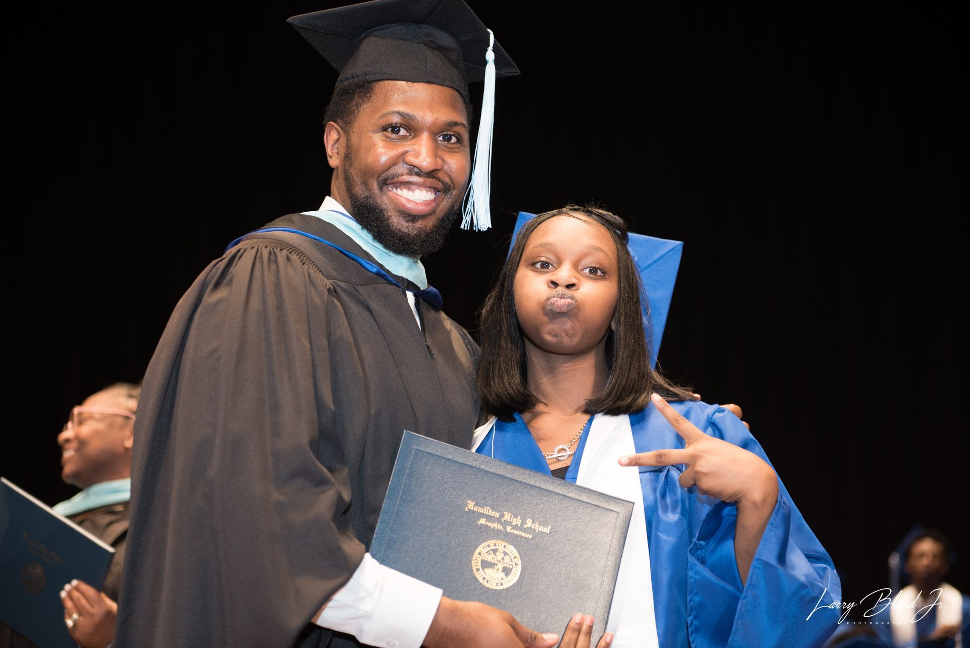 Man in graduation gown smiles with woman in blue gown making a peace sign, both holding diplomas.