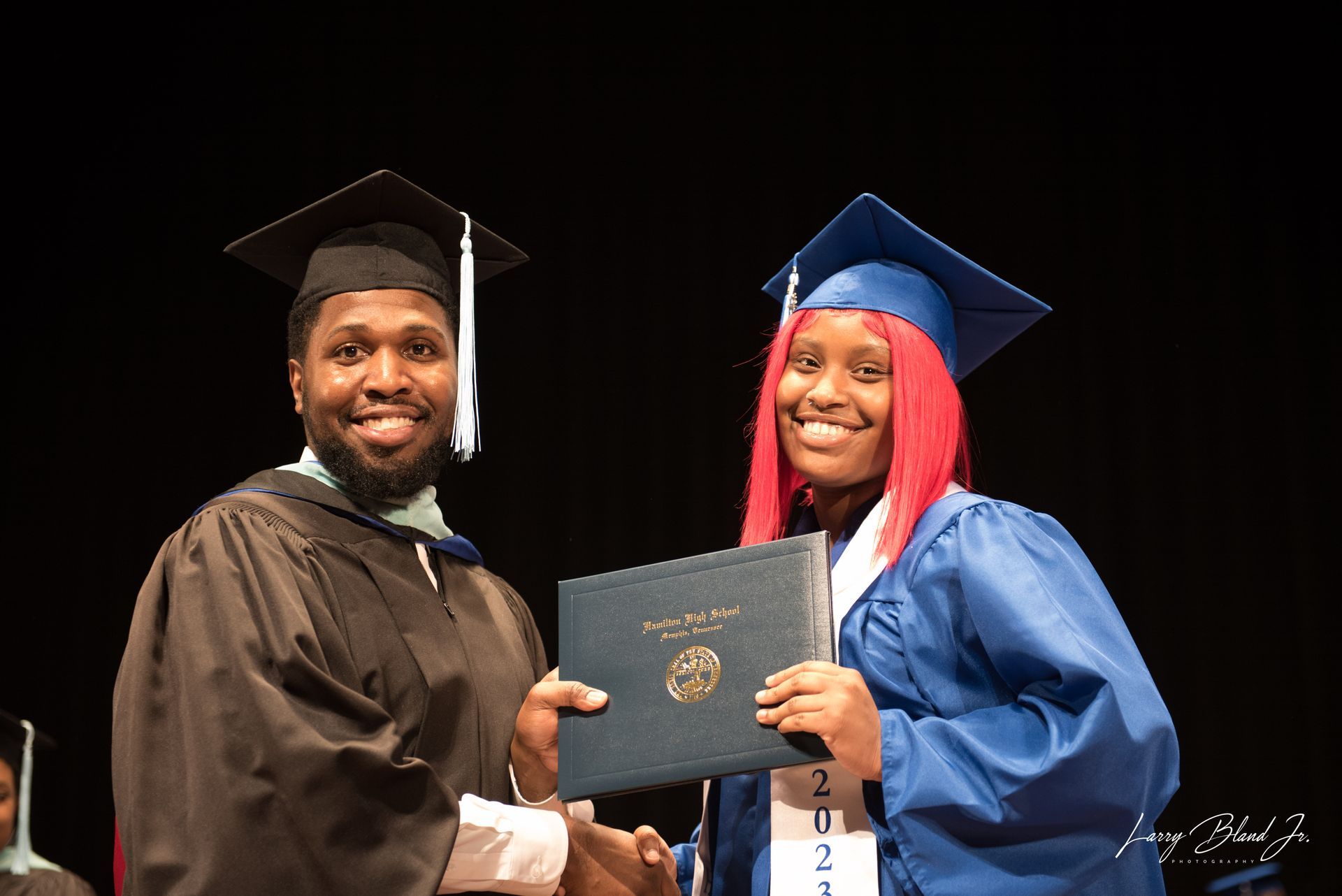 Two people in graduation attire, one handing a diploma to the other, both smiling. Stage setting.