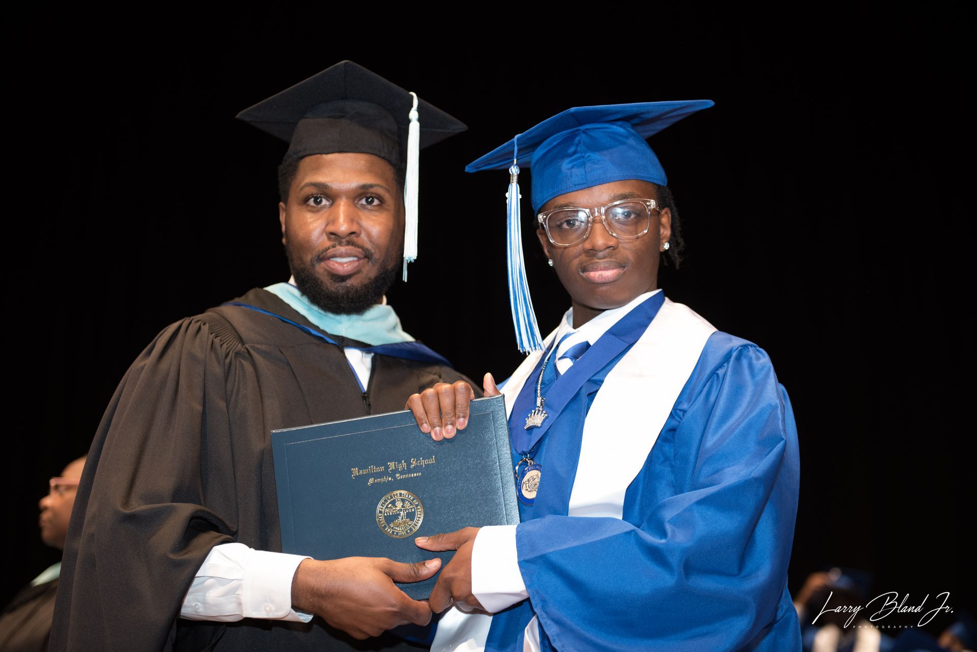 Man in black graduation gown presents diploma to person in blue gown at graduation.