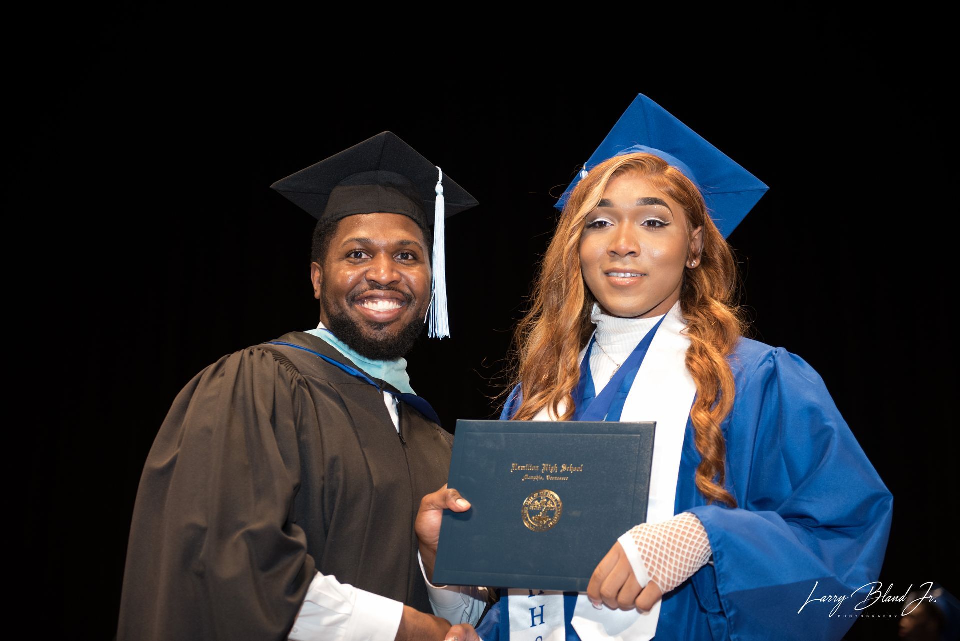 Man in robe handing a diploma to a person in a blue cap and gown. Both are smiling.