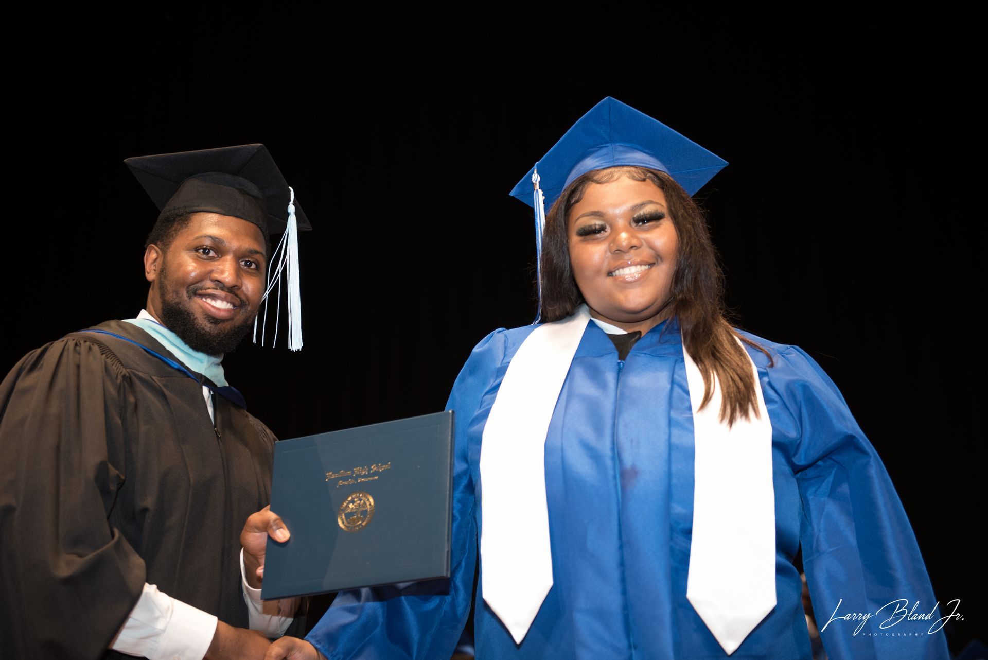 Graduation ceremony: person in blue gown receives diploma from robed person, both smiling.