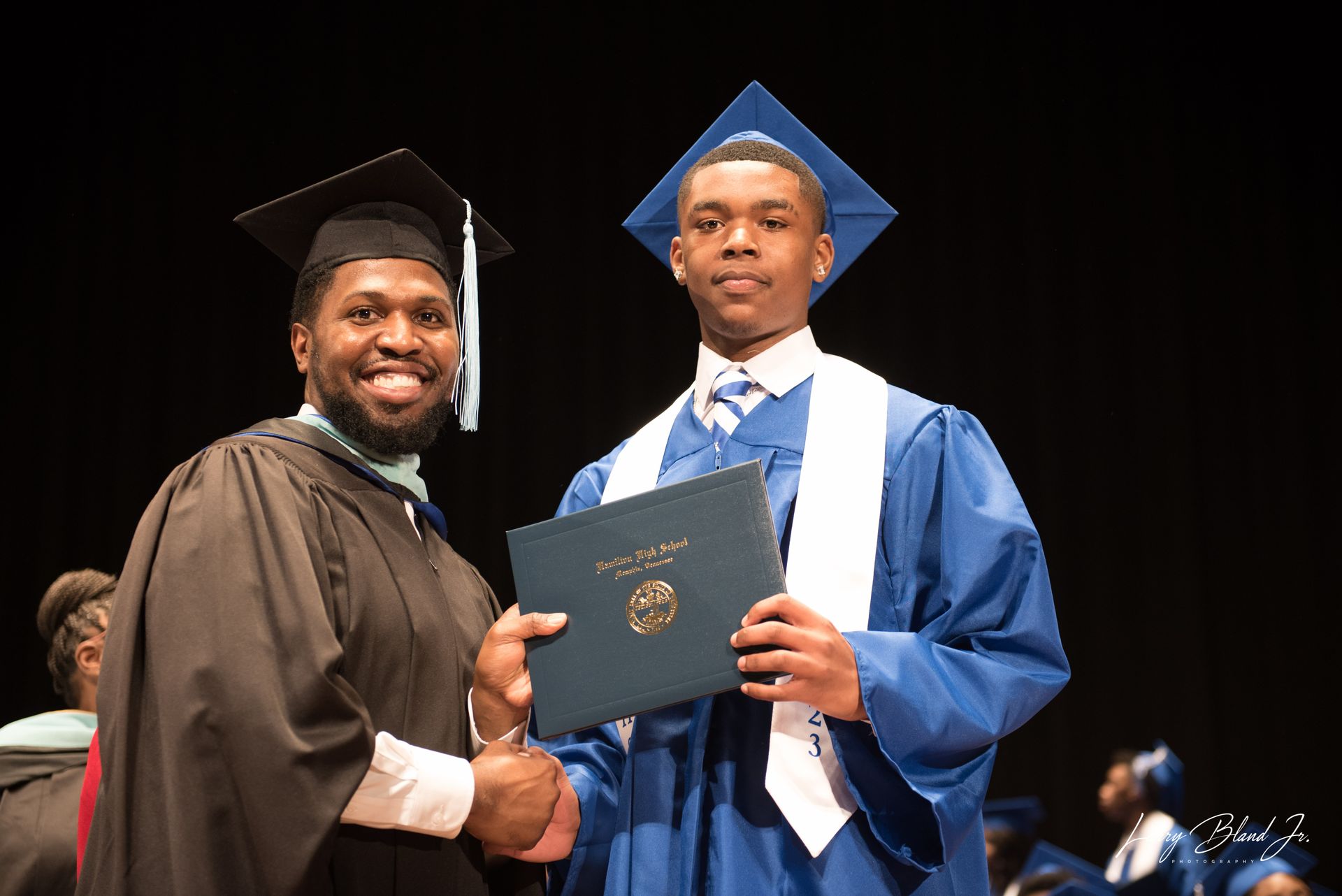 Man in academic gown hands diploma to student in blue graduation attire.