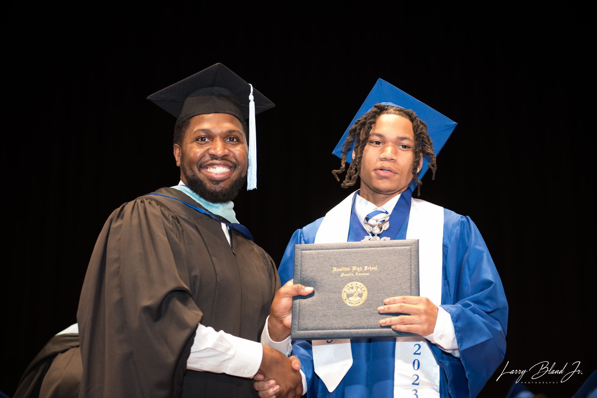 Man in cap and gown shakes hands with person in blue graduation attire, holding a diploma. Black backdrop.