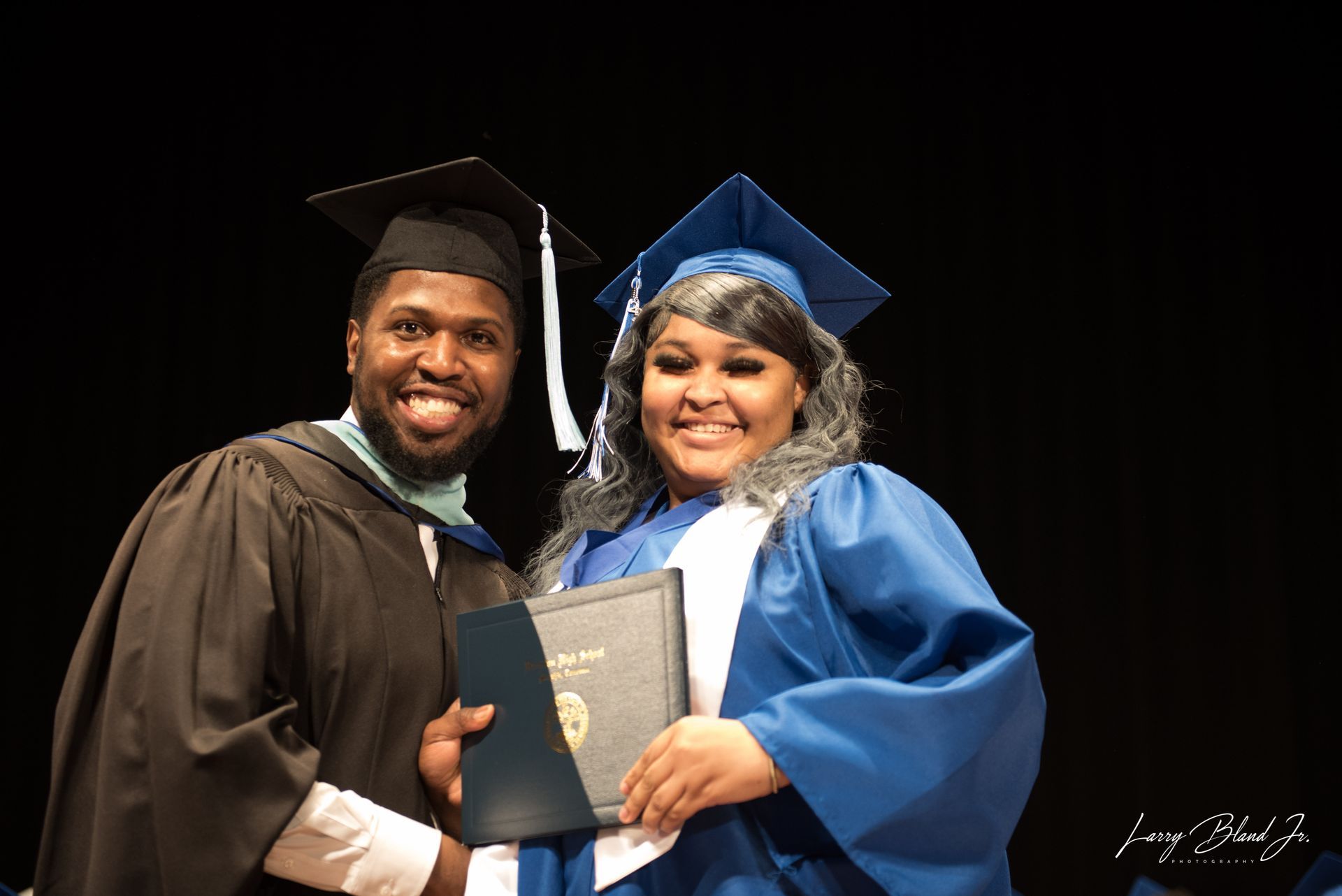 Two people in graduation attire, smiling and holding a diploma, against a dark background.