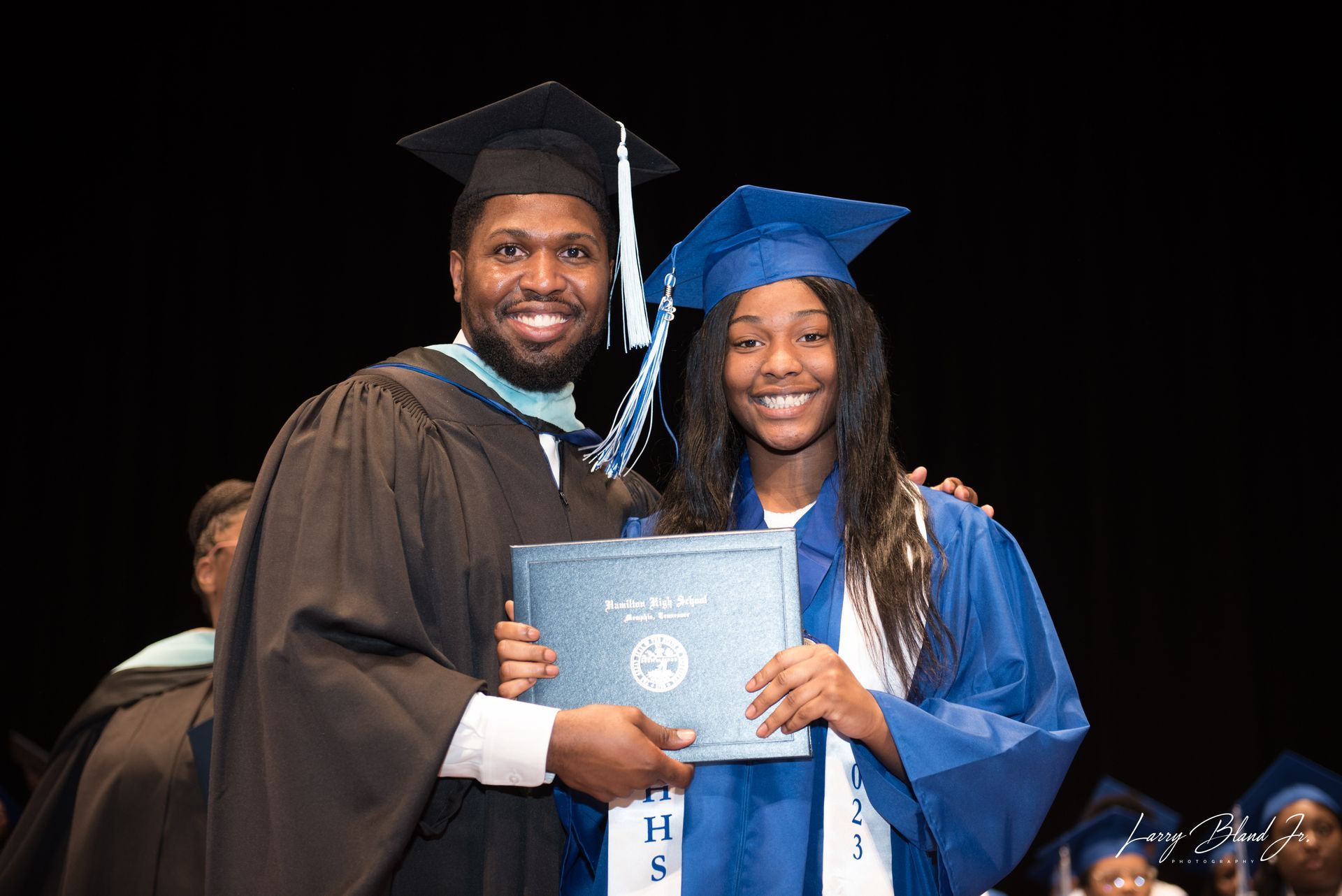 Man in graduation gown handing a diploma to a person in a blue cap and gown on stage.