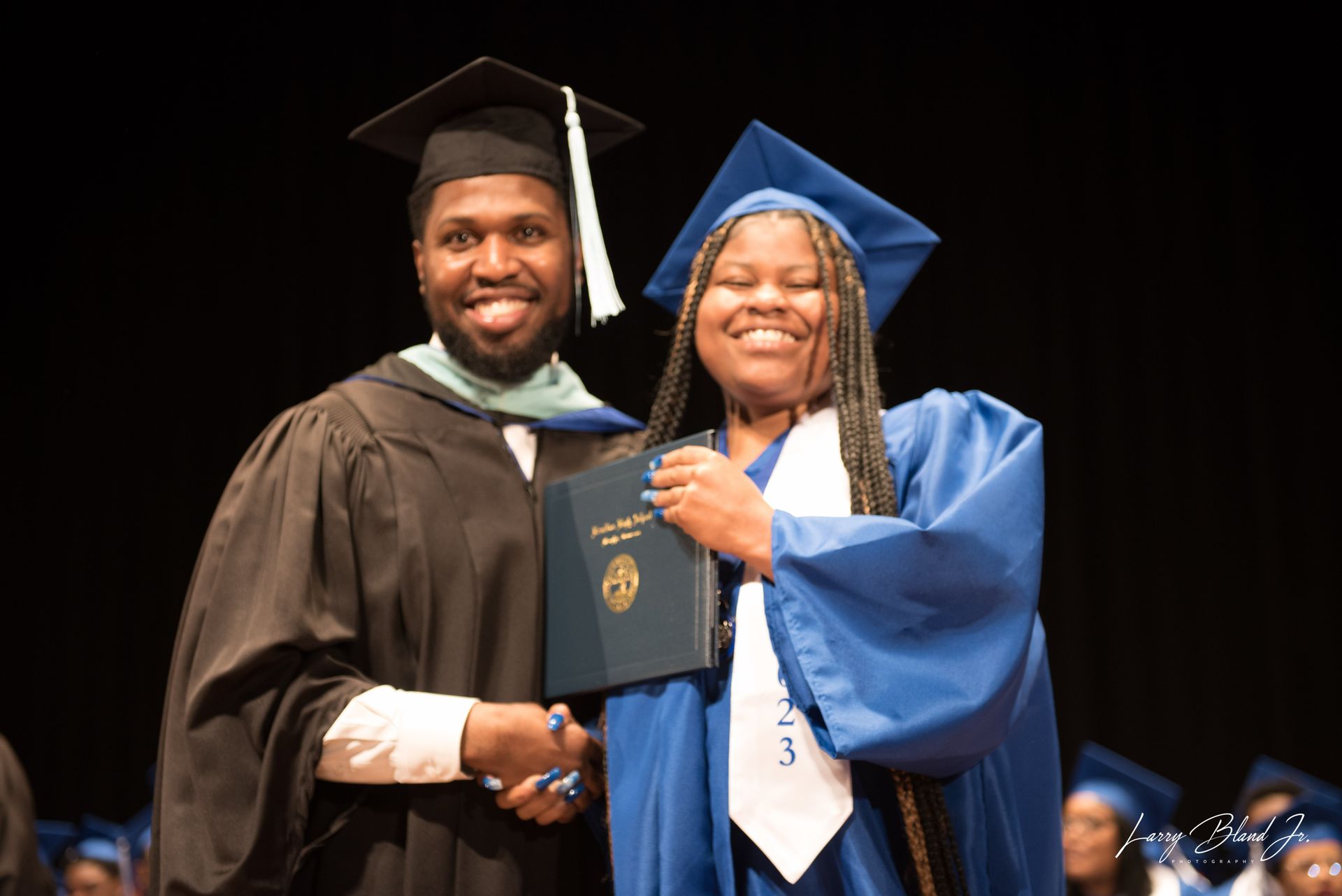 Man in black graduation gown shakes hands with a woman in blue gown holding a diploma. Both are smiling.