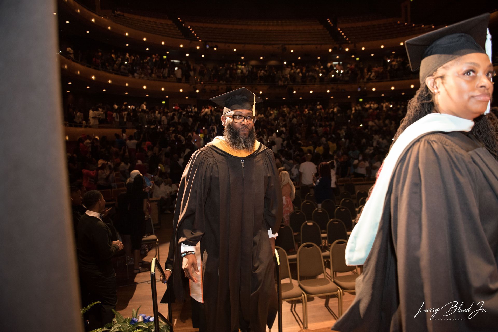 Graduates in black gowns and mortarboards walk across a stage in a packed auditorium.
