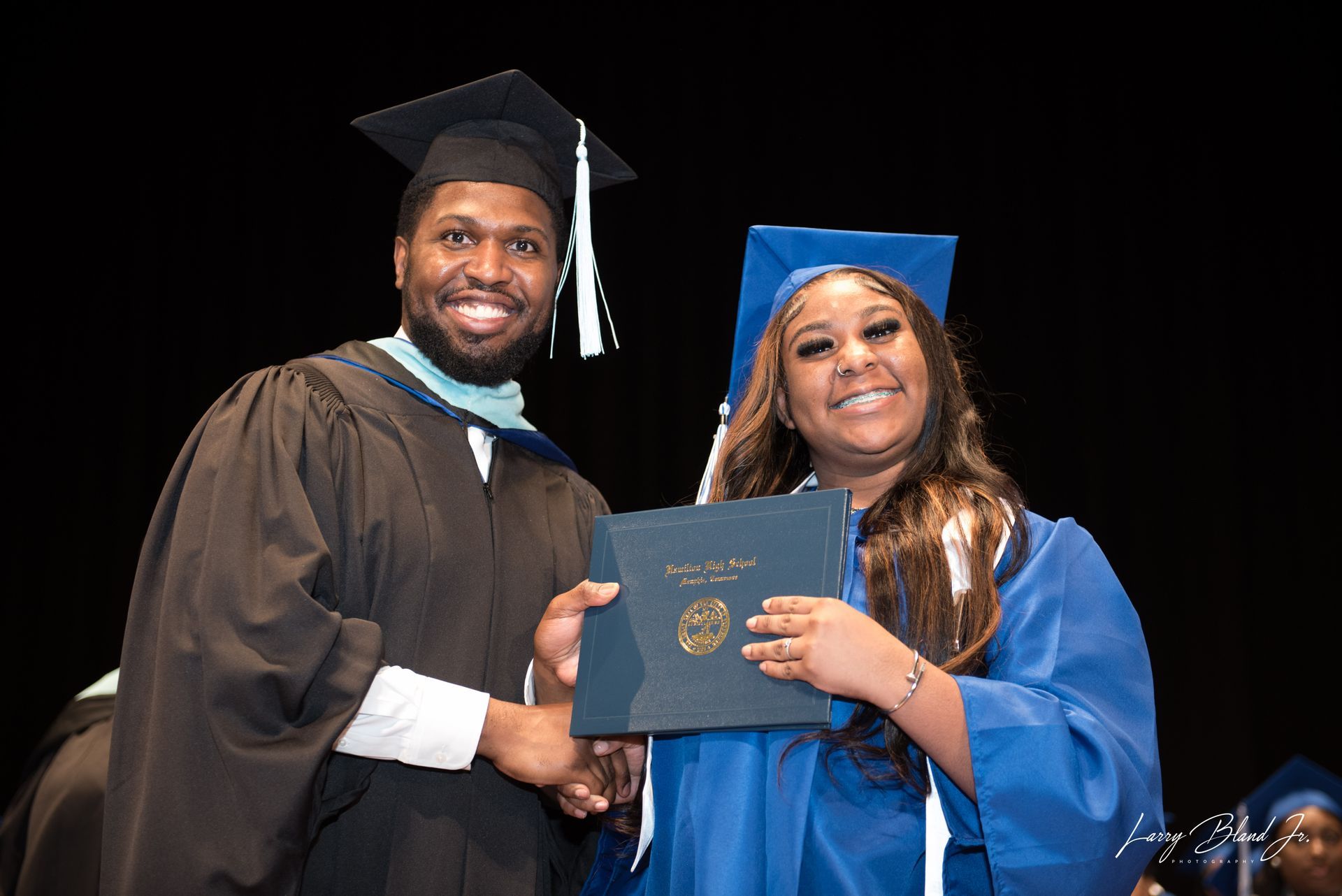 Man in graduation robe handing a diploma to a person in a blue graduation gown on stage.