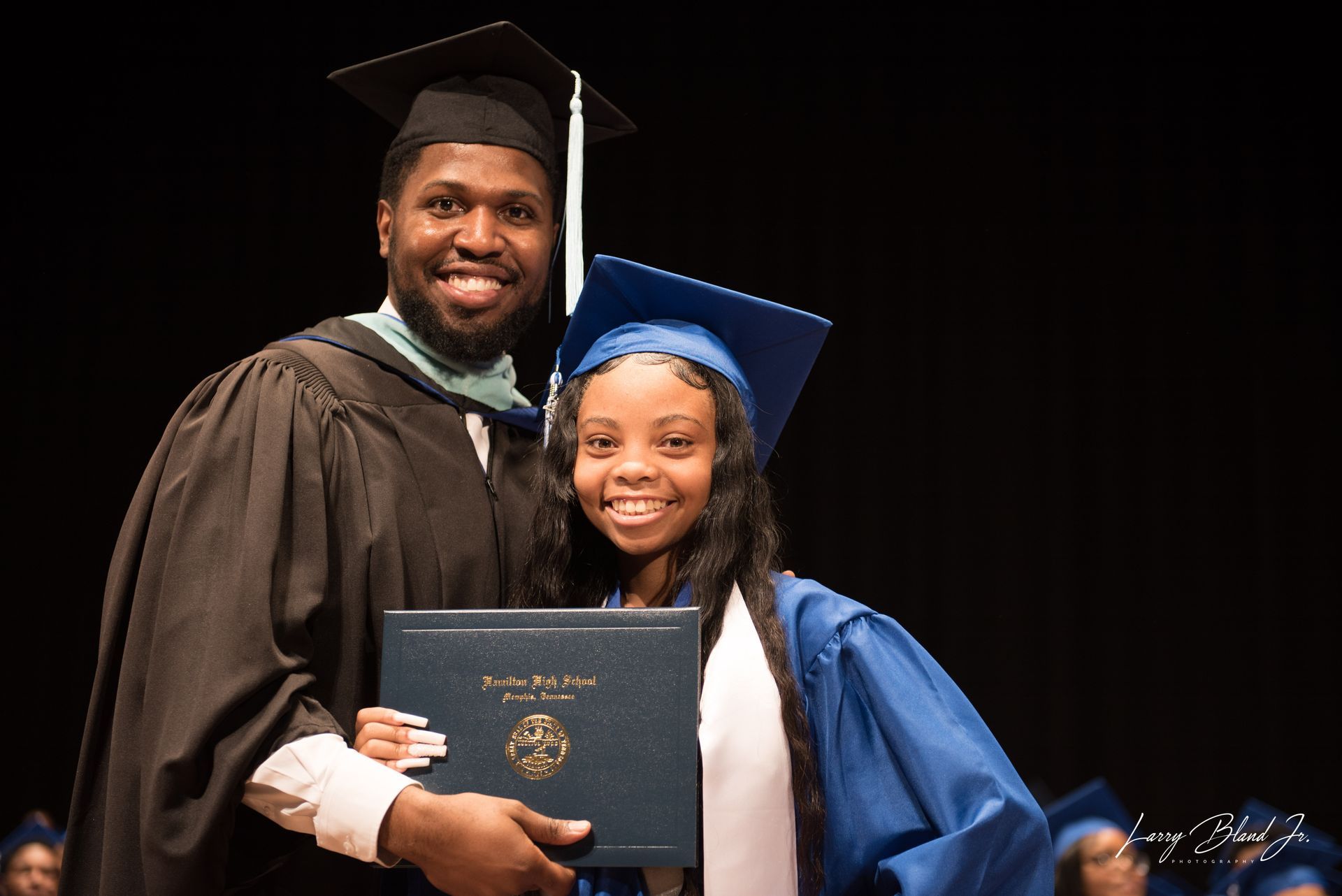 Man in cap and gown embraces a smiling graduate in blue, holding a diploma, on stage.