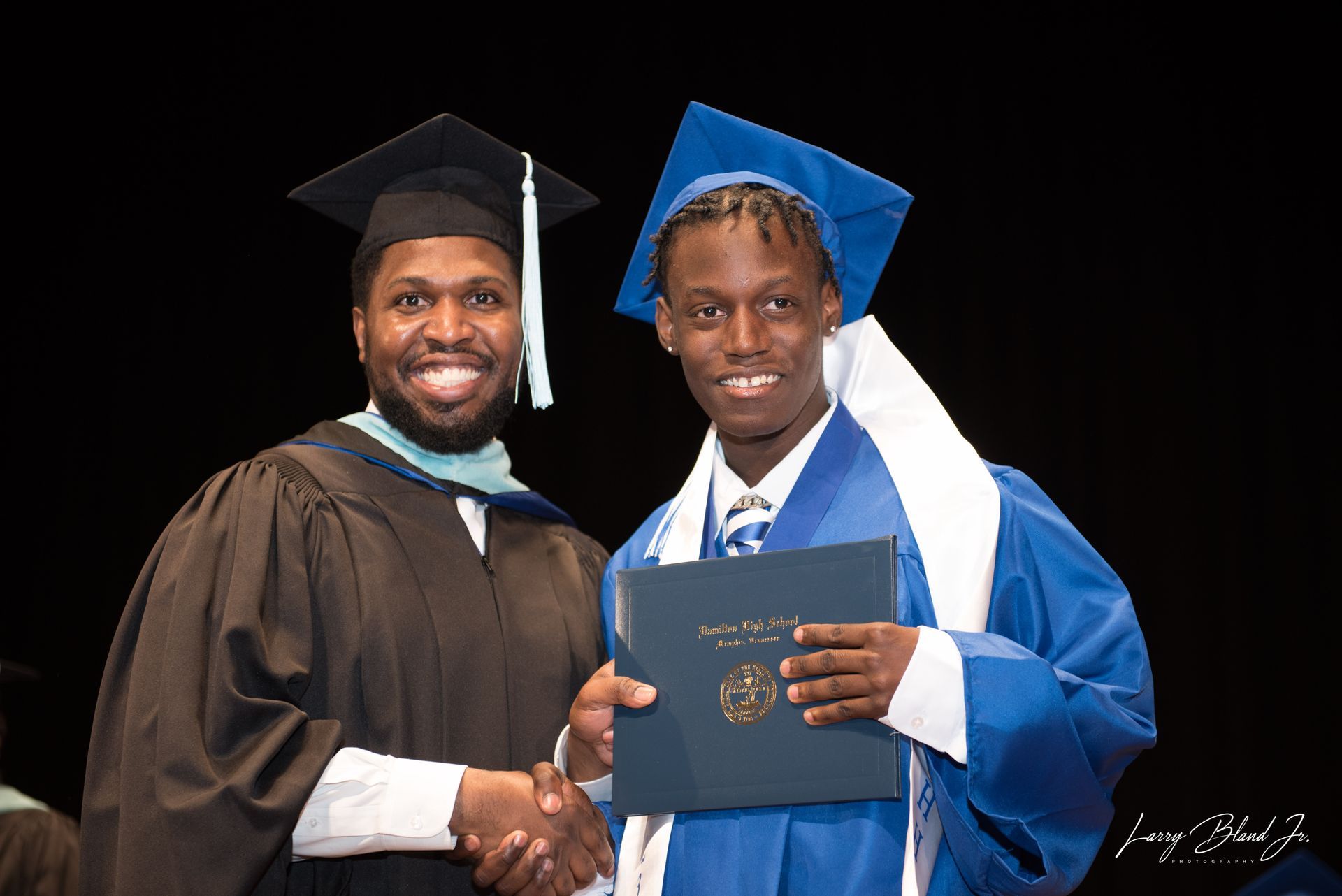 Man in cap and gown shakes hands with graduate holding diploma, both smiling.
