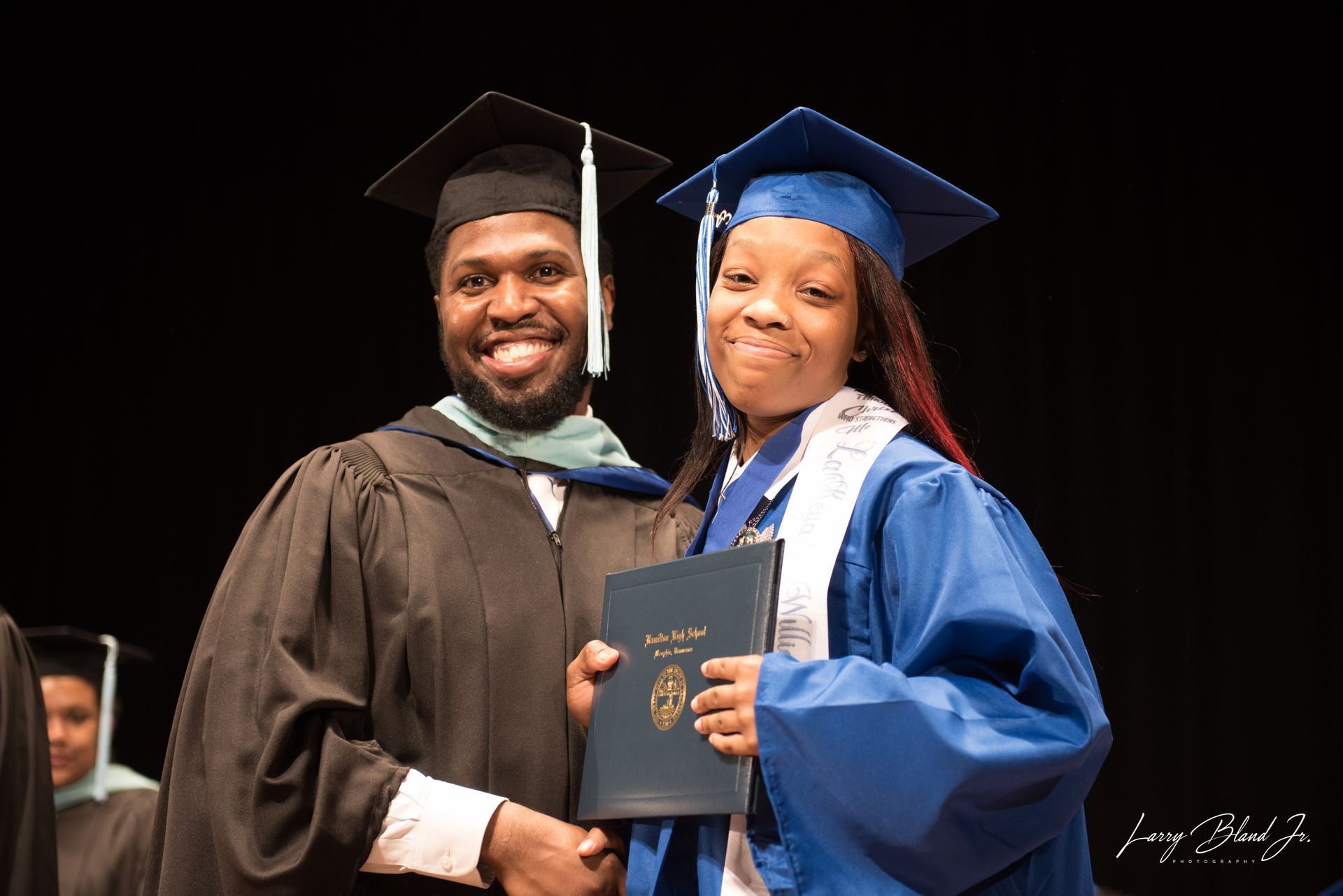 Two people in graduation gowns, one handing a diploma. The backdrop is a stage.