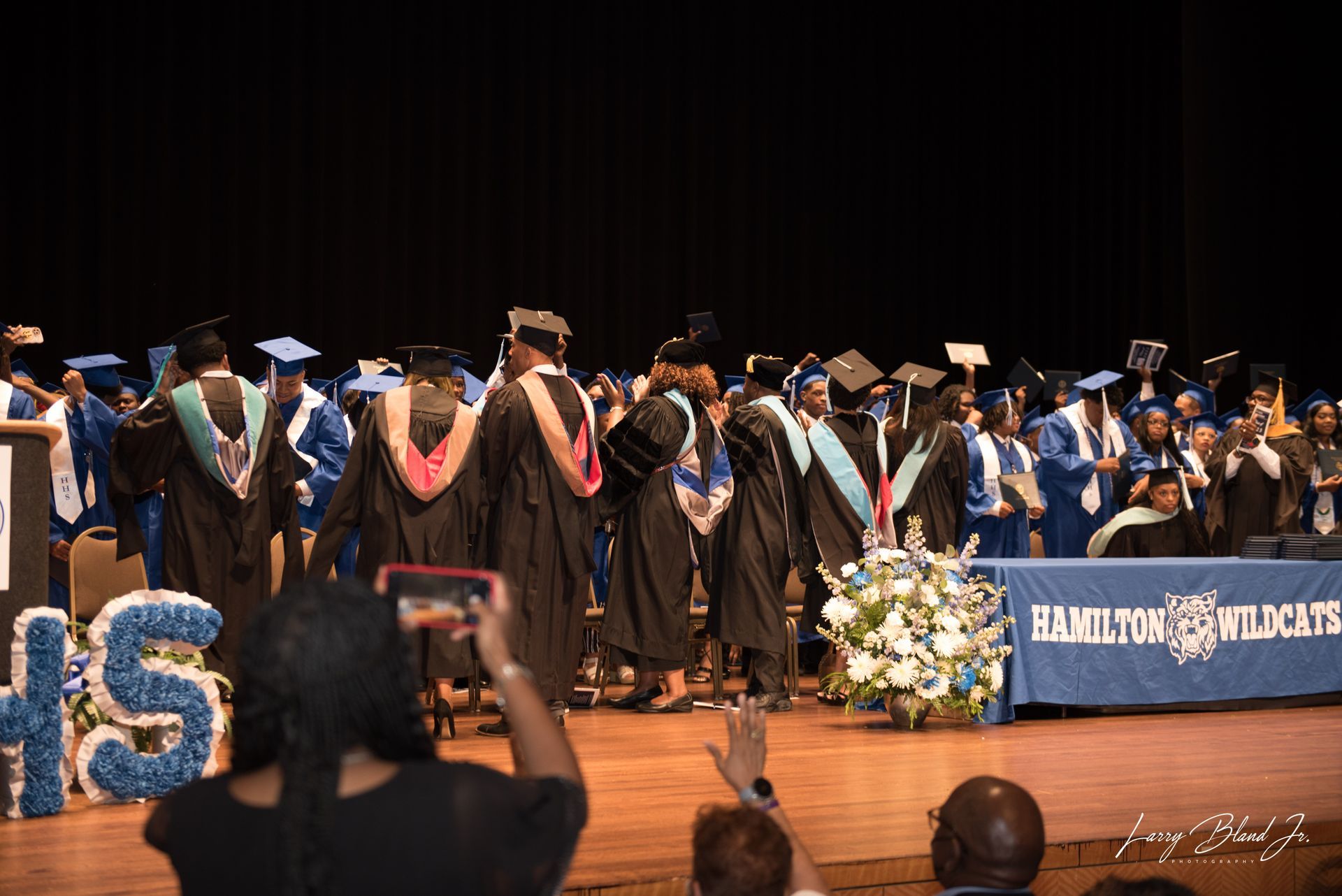 Graduates in blue and black gowns on stage at Hamilton High School graduation.