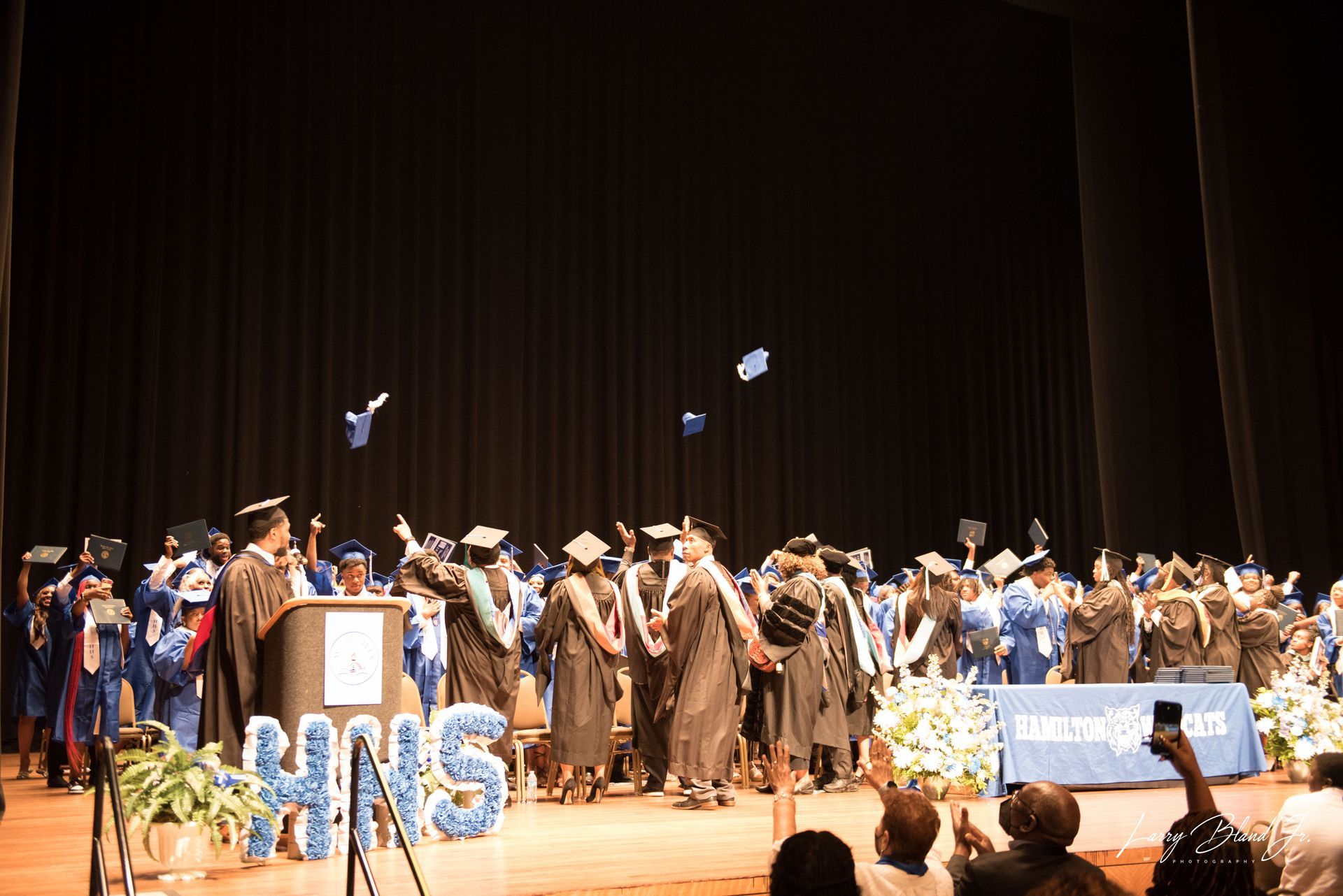 Graduates in blue and black gowns throw caps in the air on a stage with a 