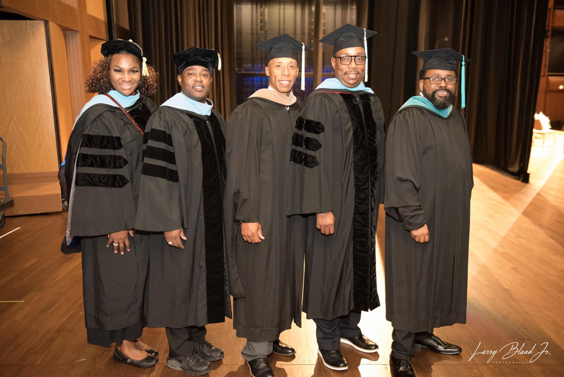 Five people in graduation gowns pose in a wood-paneled room.