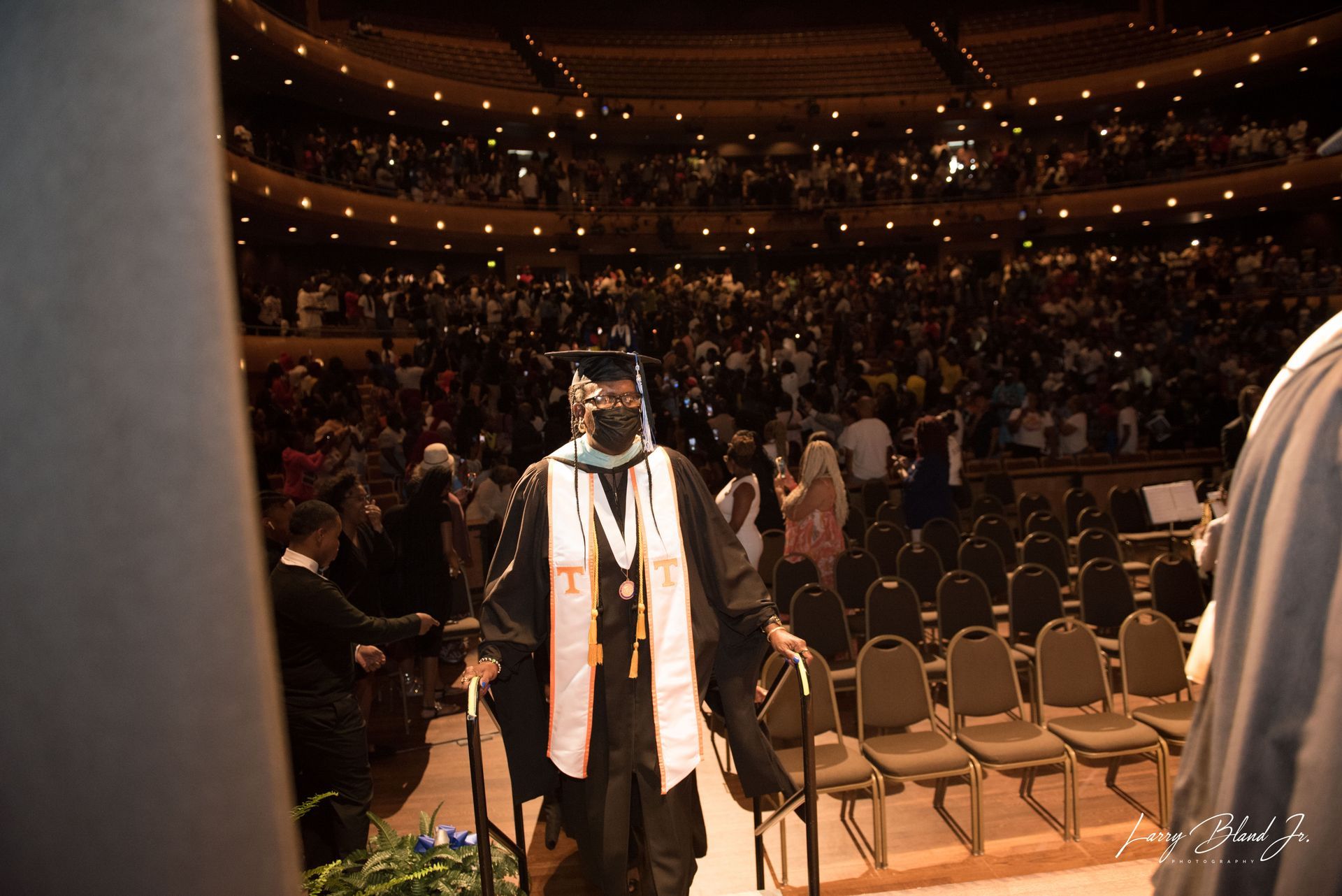 Person in graduation attire on stage, walking toward the camera, in a packed auditorium.
