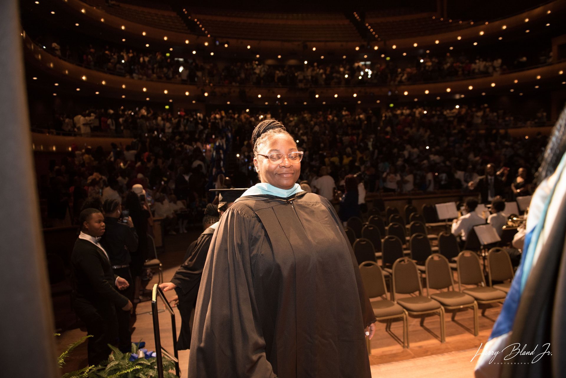 Person in graduation gown on stage, auditorium in background.