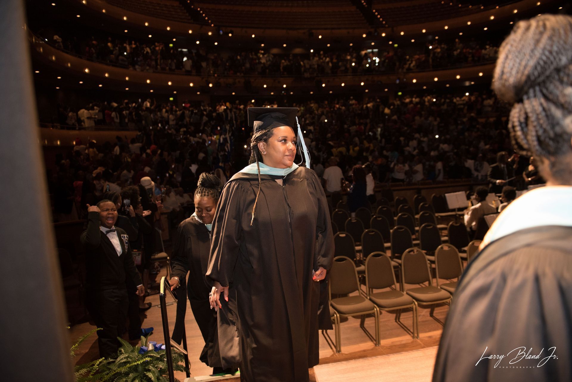 Person in graduation gown walks on stage at ceremony. Auditorium with audience visible.