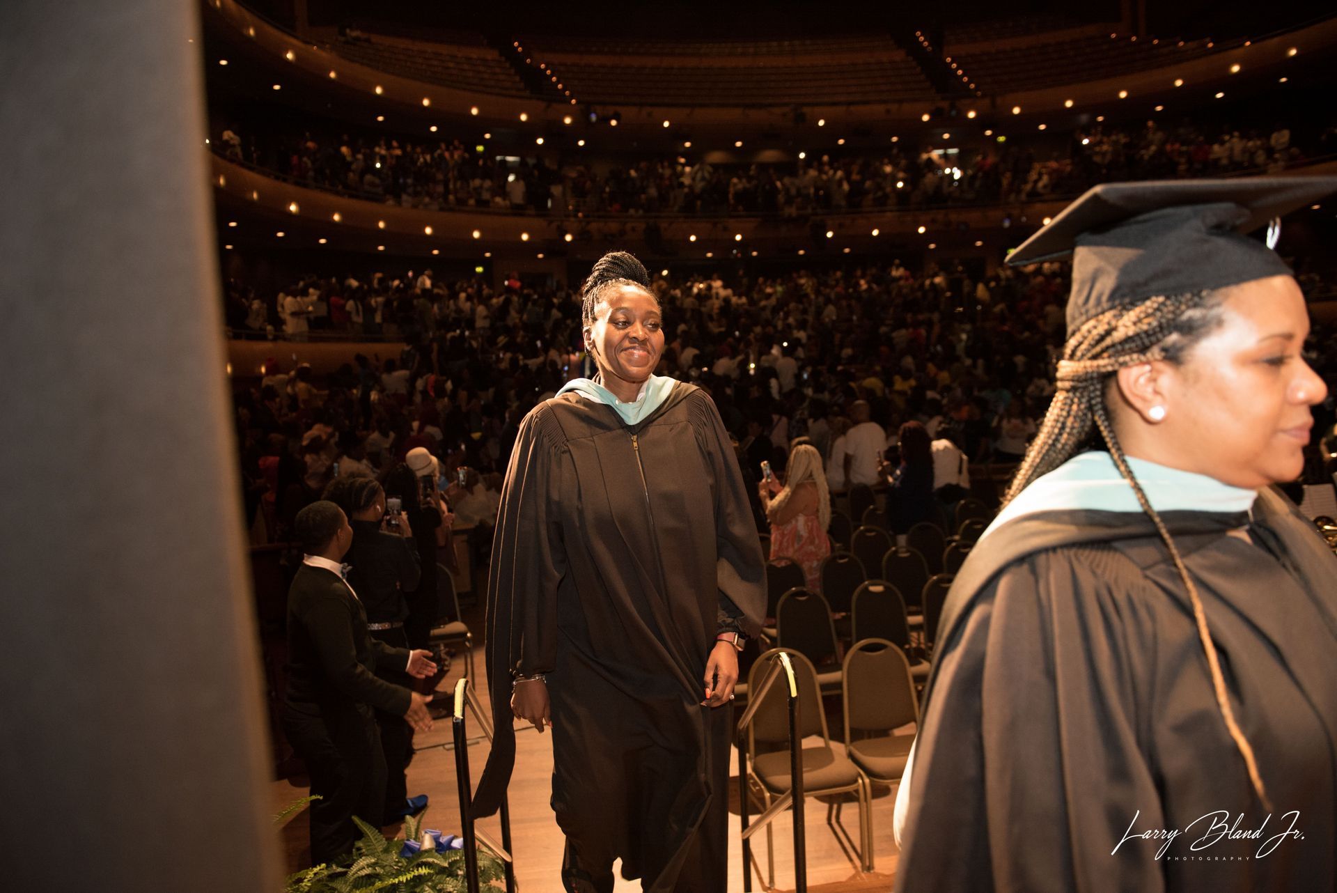 Graduation: Two graduates in black robes with stoles and caps walk on stage, smiling; large auditorium in background.