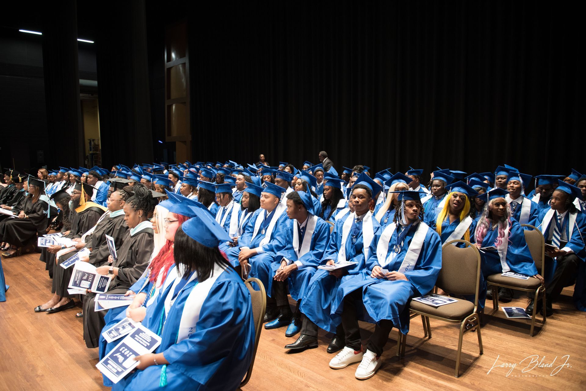 Graduation ceremony: graduates in blue gowns and caps seated in an auditorium.