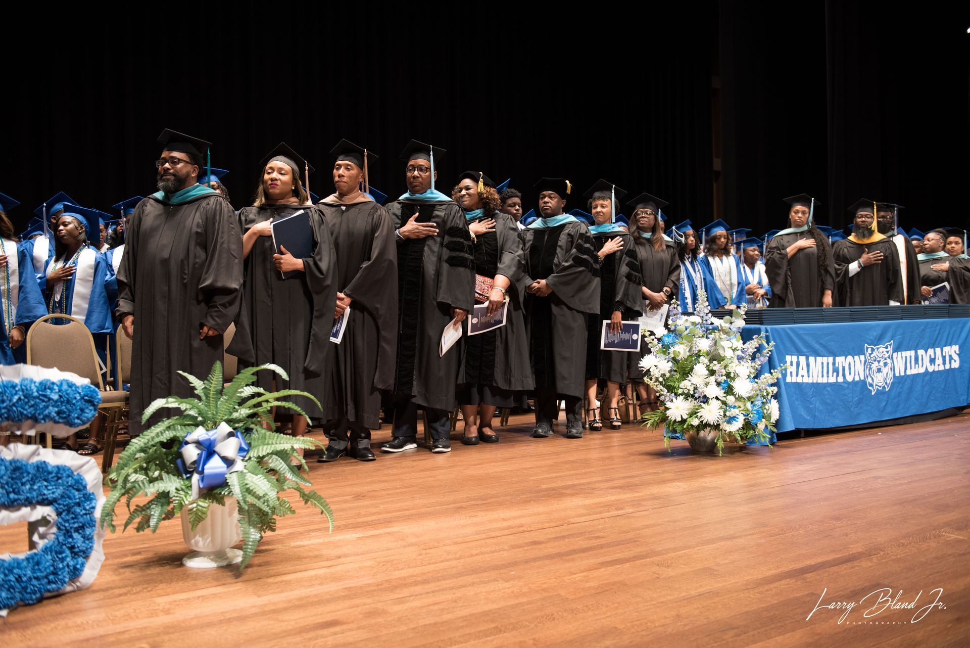 Graduates in black gowns and blue stoles stand onstage, hands over hearts, during a ceremony.