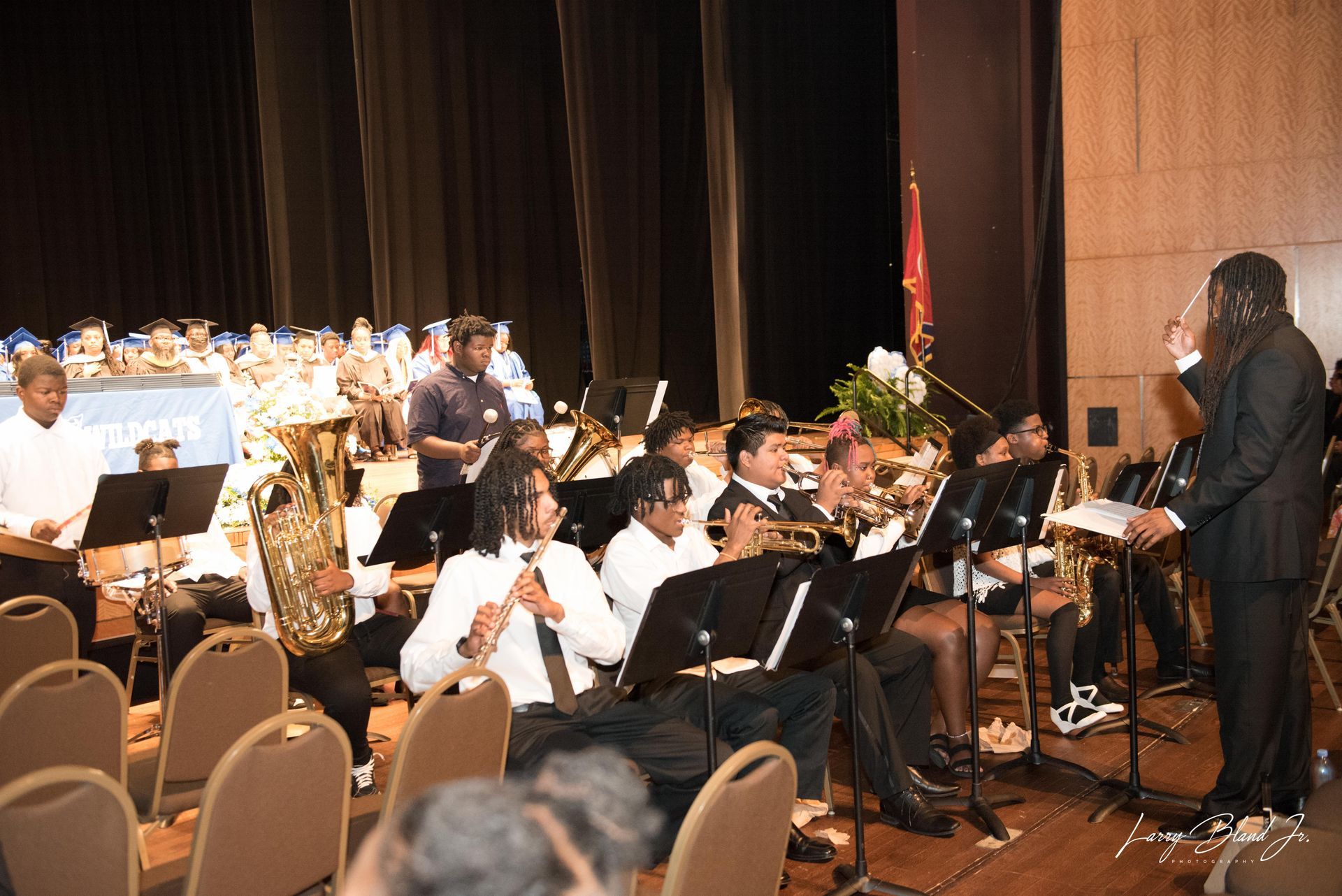 Musical ensemble performing on stage; conductor, brass, and woodwind instruments.