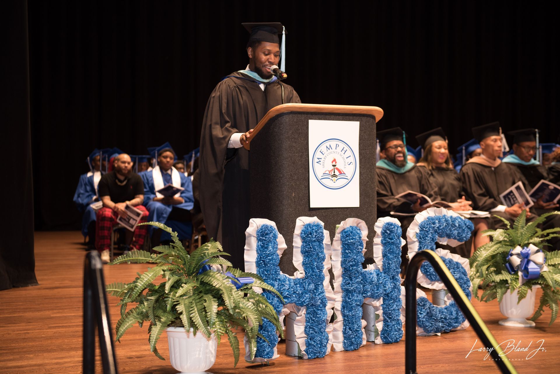 A graduate in a cap and gown giving a speech at a podium on a stage during a ceremony.