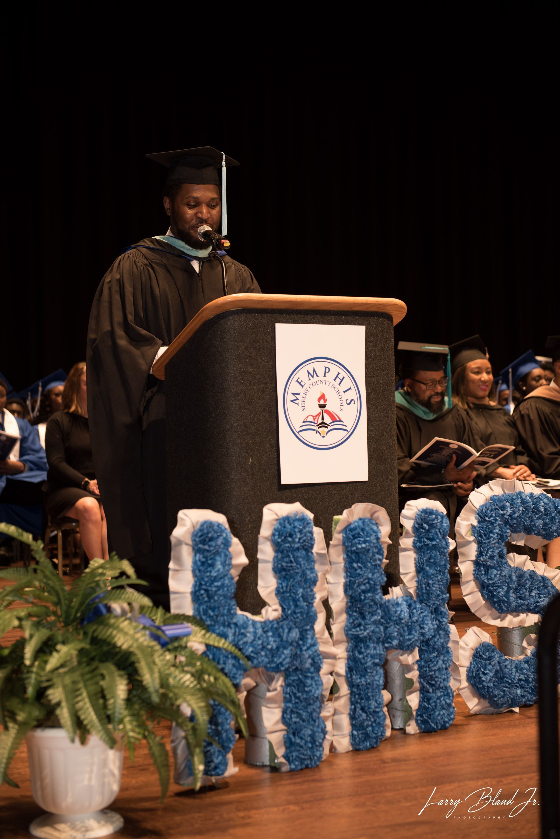 A graduate in cap and gown speaks at a podium with school logo, 