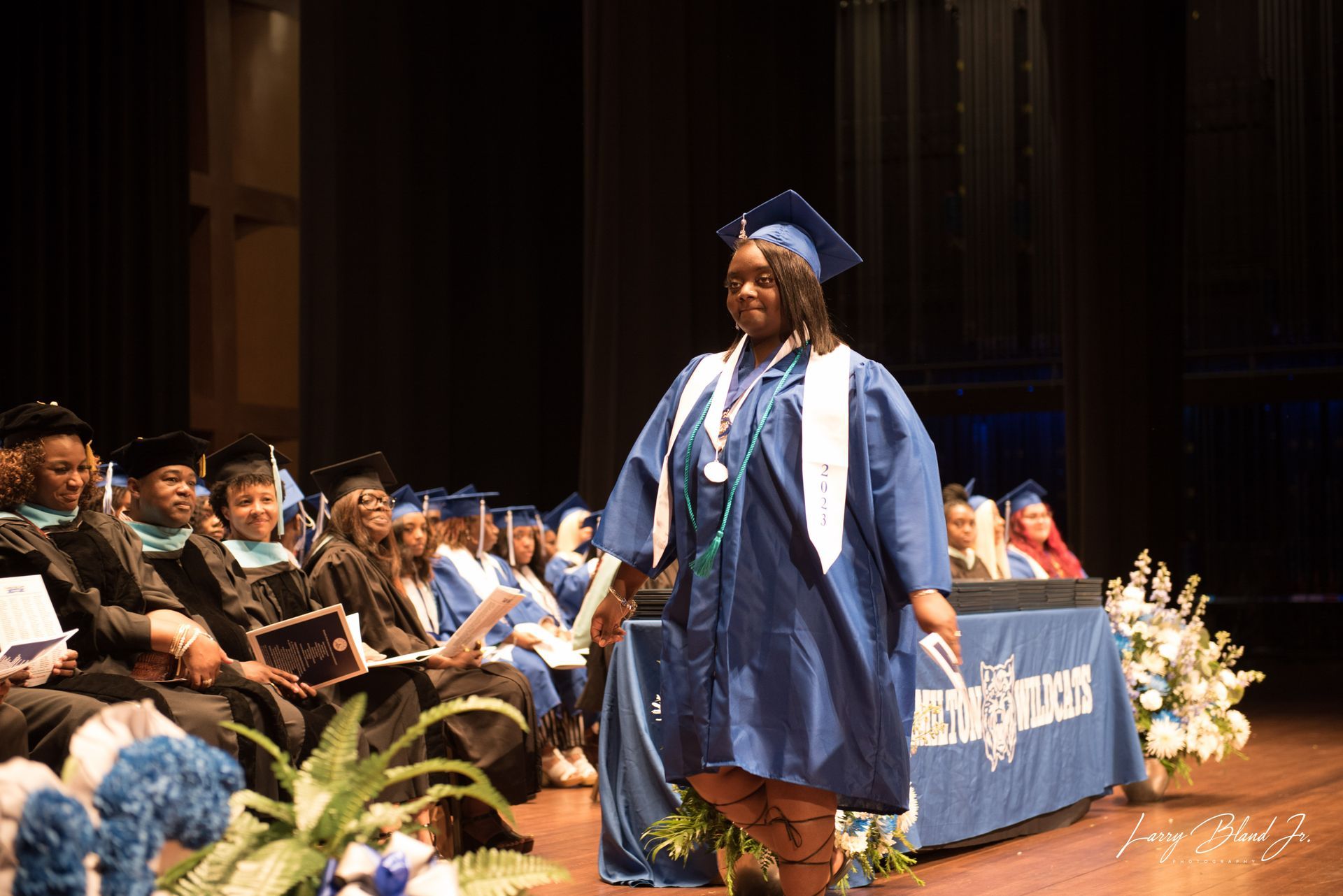 Graduate in blue gown walking across a stage. Others in gowns seated in background.