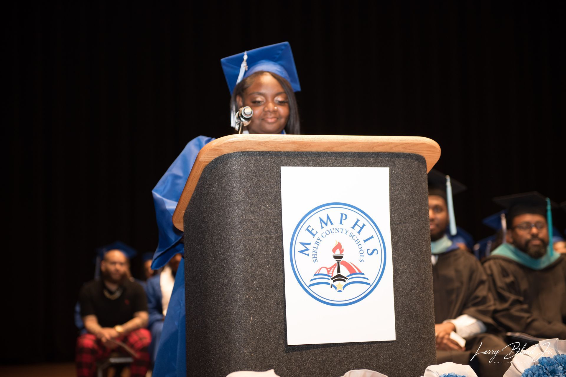 Person in graduation attire speaking at podium with a Memphis University logo.