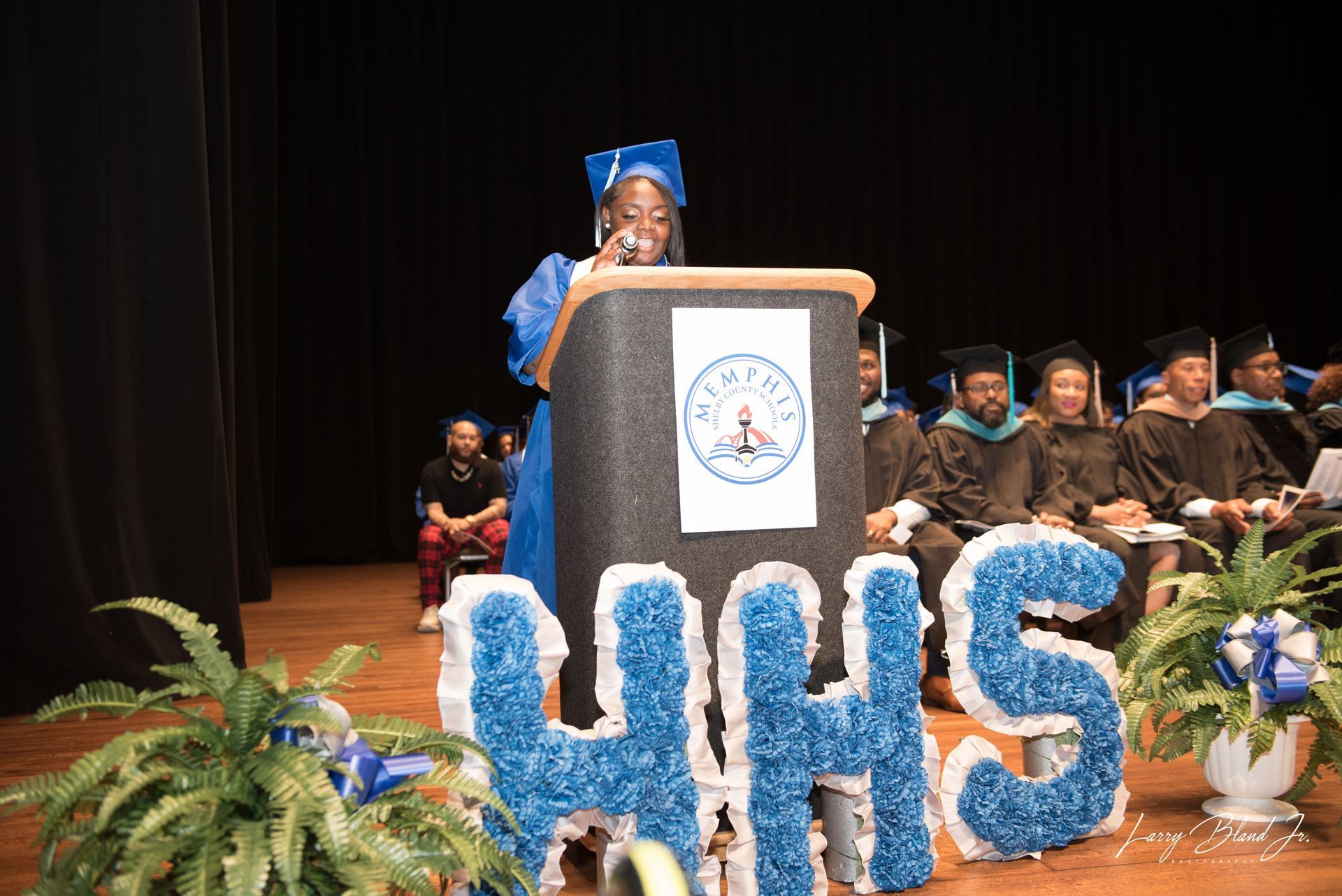 Graduation ceremony: person in blue cap and gown speaks at podium, 