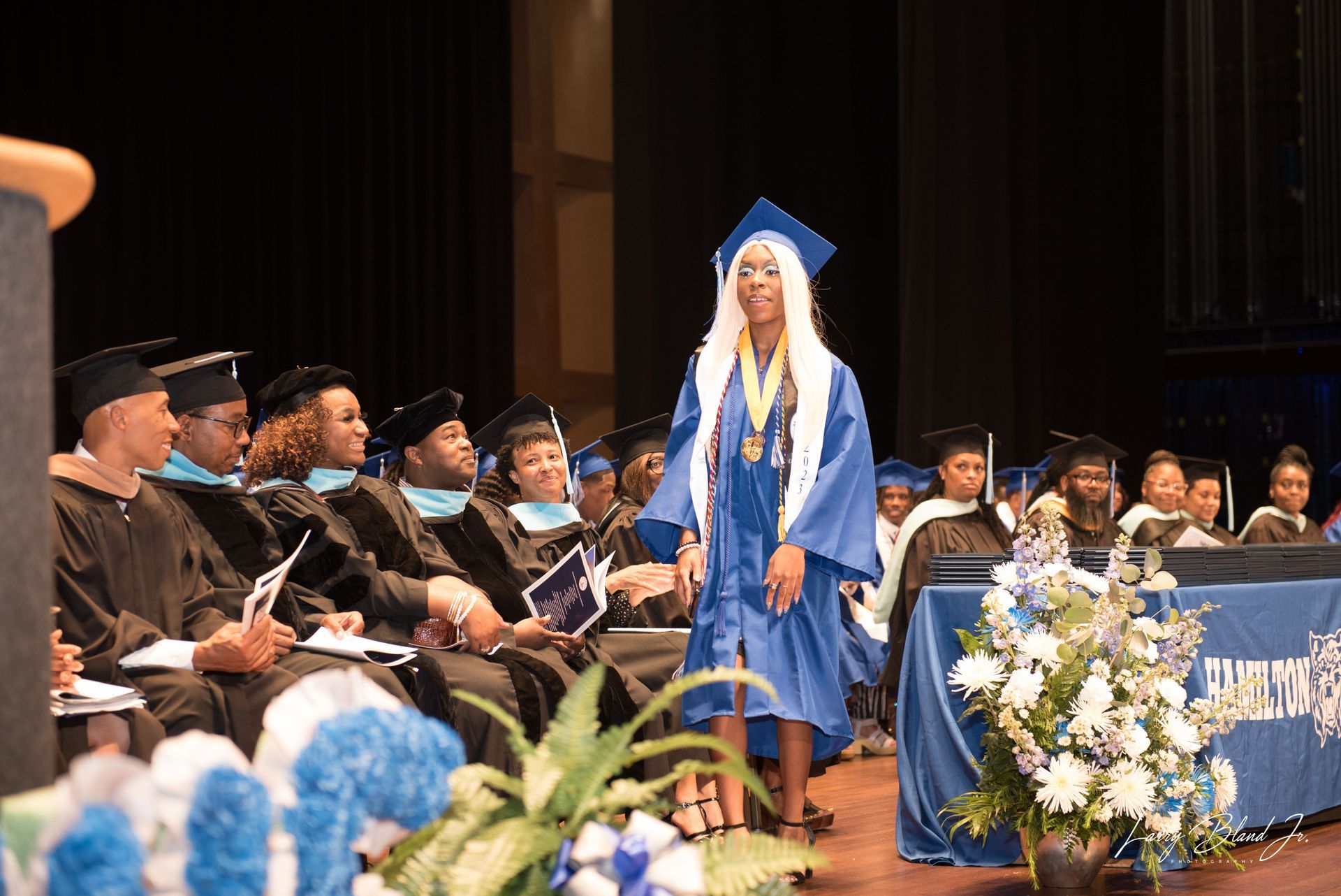 Student in blue graduation gown walks across stage, ceremony setting with audience.