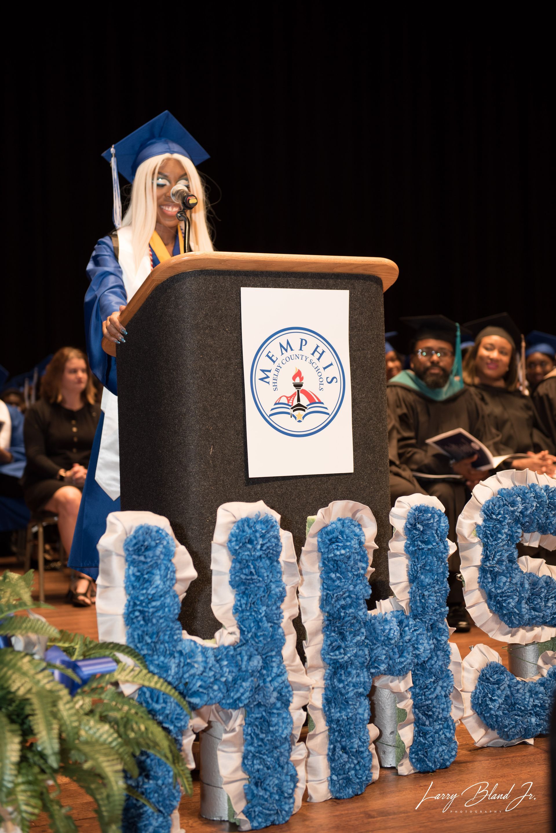 Graduate giving speech at podium. Blue and white decorations.  Ceremony hall setting.