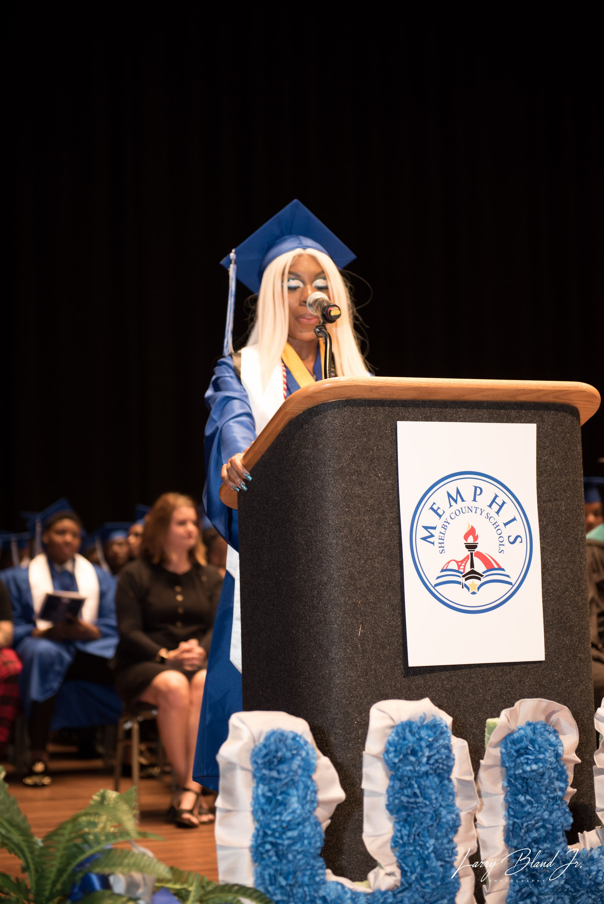 Student in graduation gown giving a speech at a podium with the word 