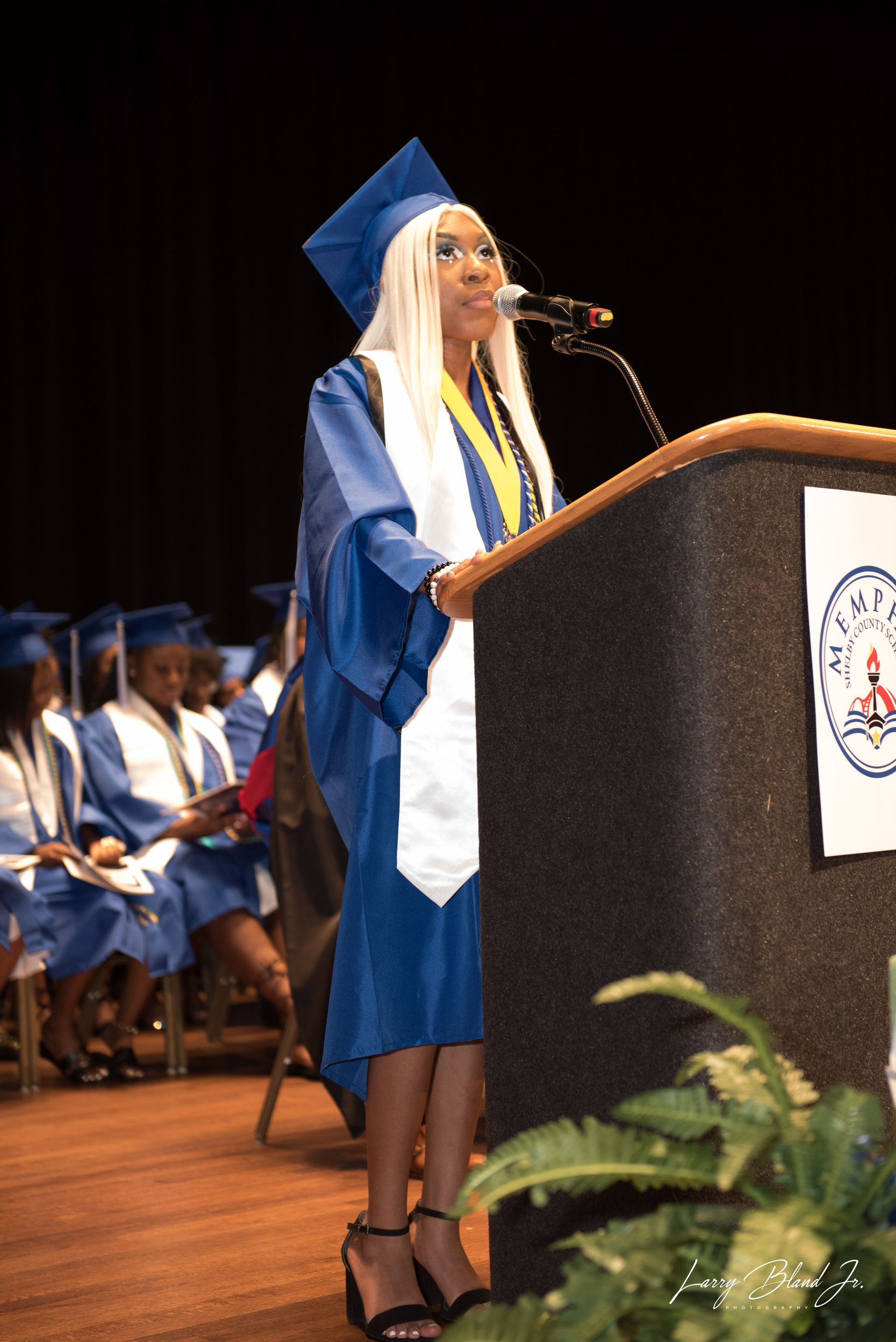 Graduate in blue gown, white stole, blonde hair speaks at podium.