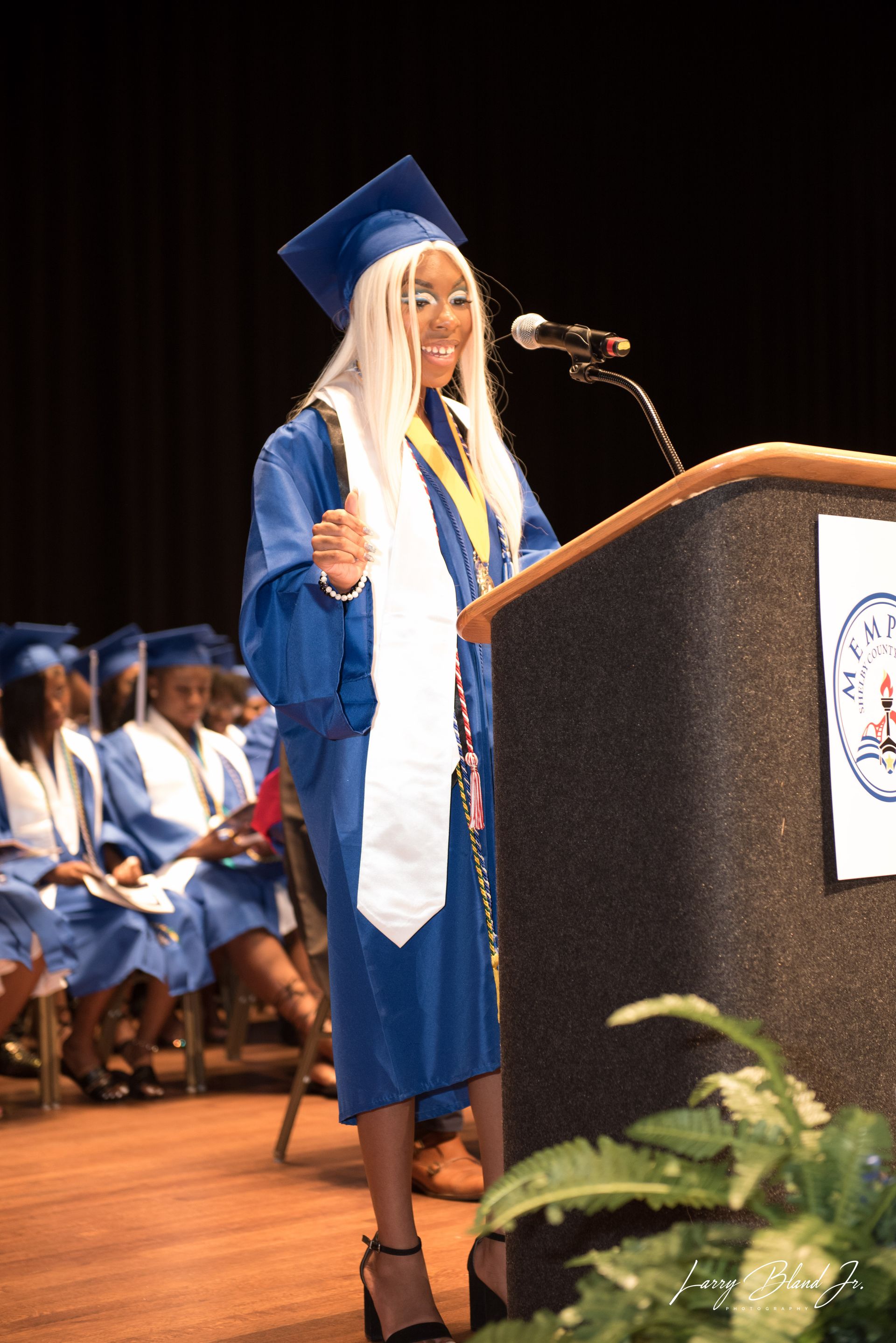 Graduation speech: Person in blue cap and gown at podium, smiling, gesturing, with audience in background.
