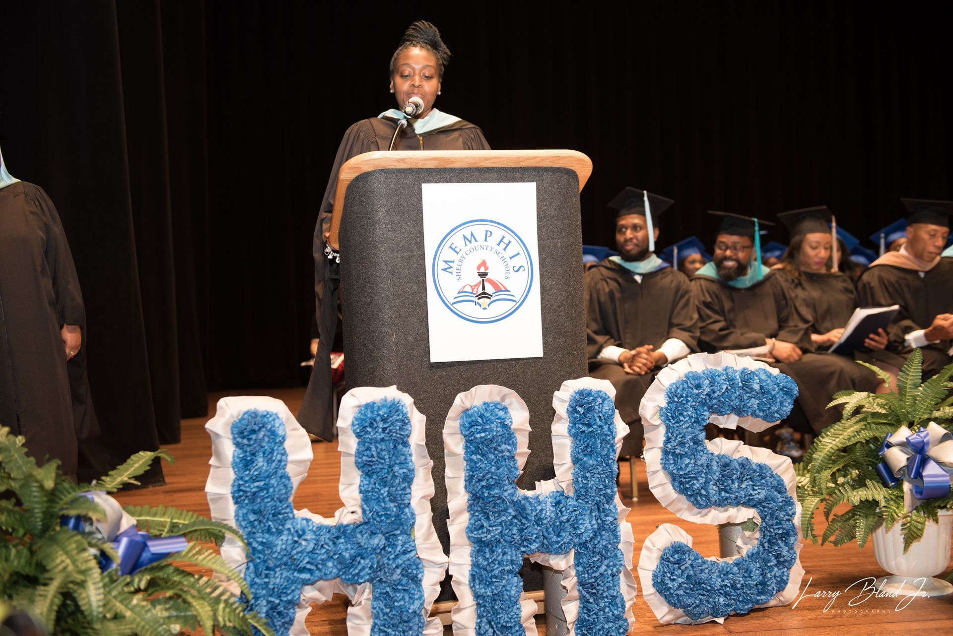 A person in a graduation gown speaks at a podium with 