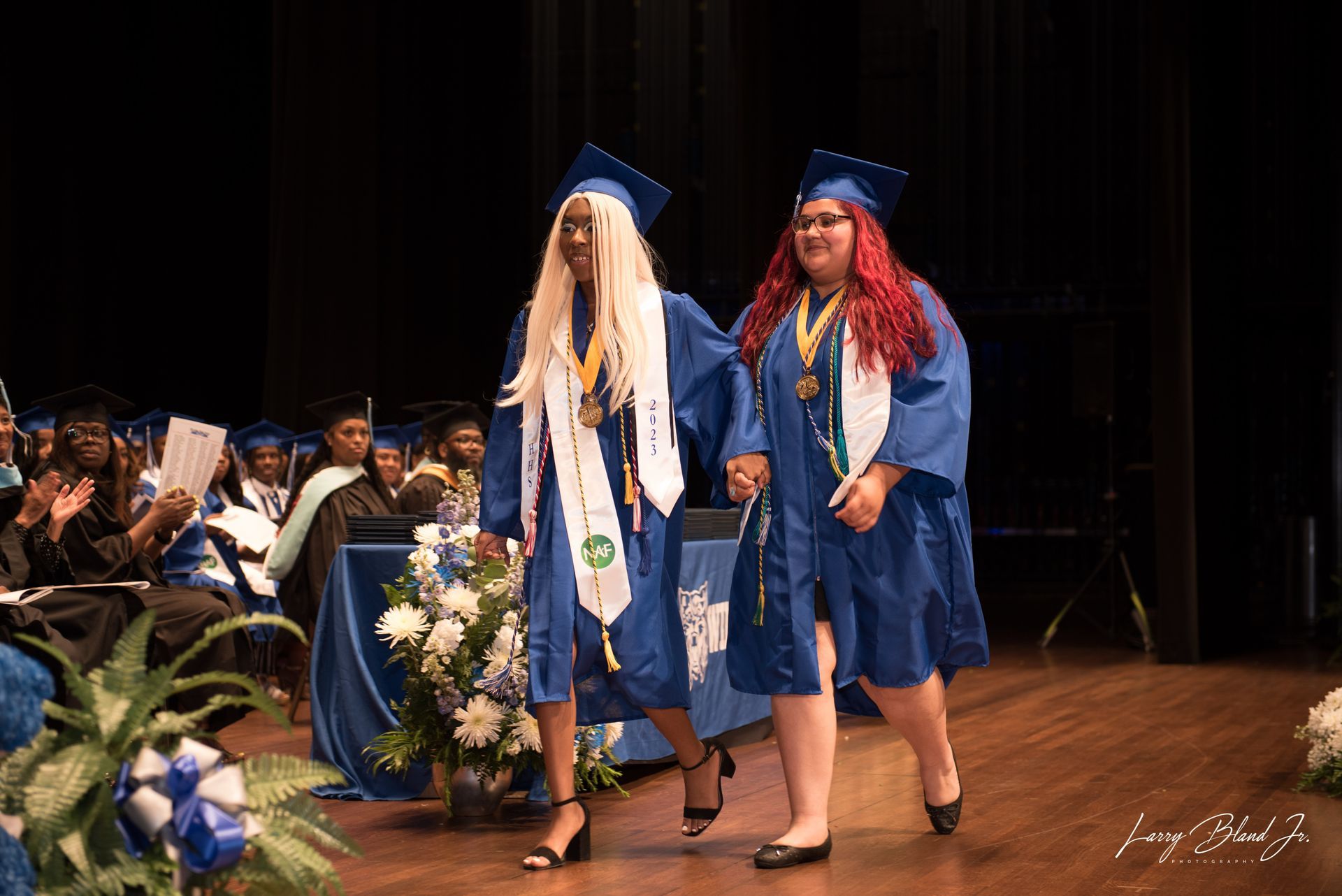 Two graduates in blue gowns and caps walk on stage, holding hands, at graduation.