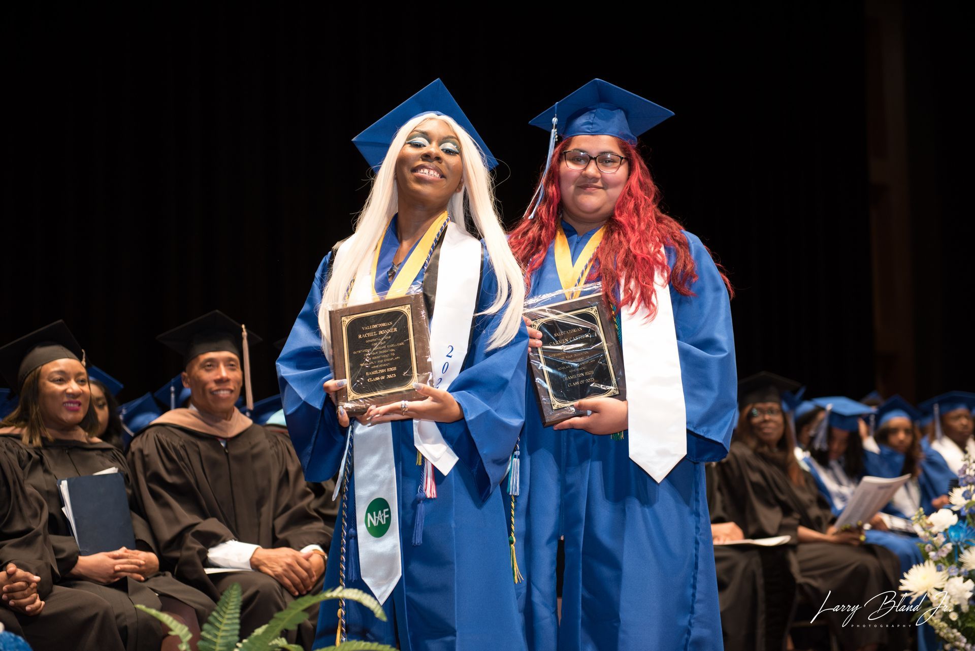 Two graduates in blue gowns and caps holding awards on stage.