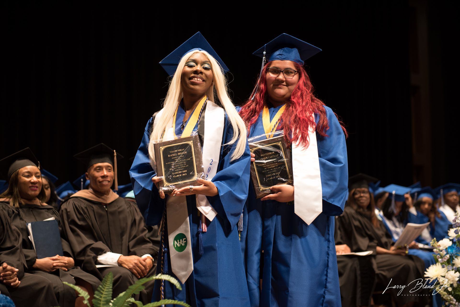 Two graduates in blue gowns and caps hold awards, smiling, on stage.