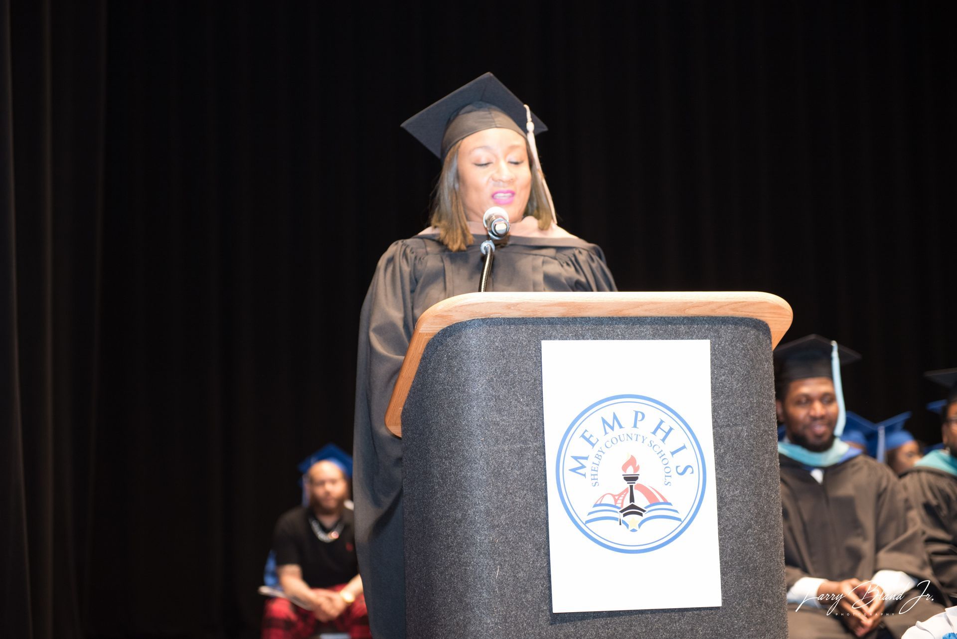 A graduate in cap and gown speaks at a podium with the university logo. Other graduates sit in the background.