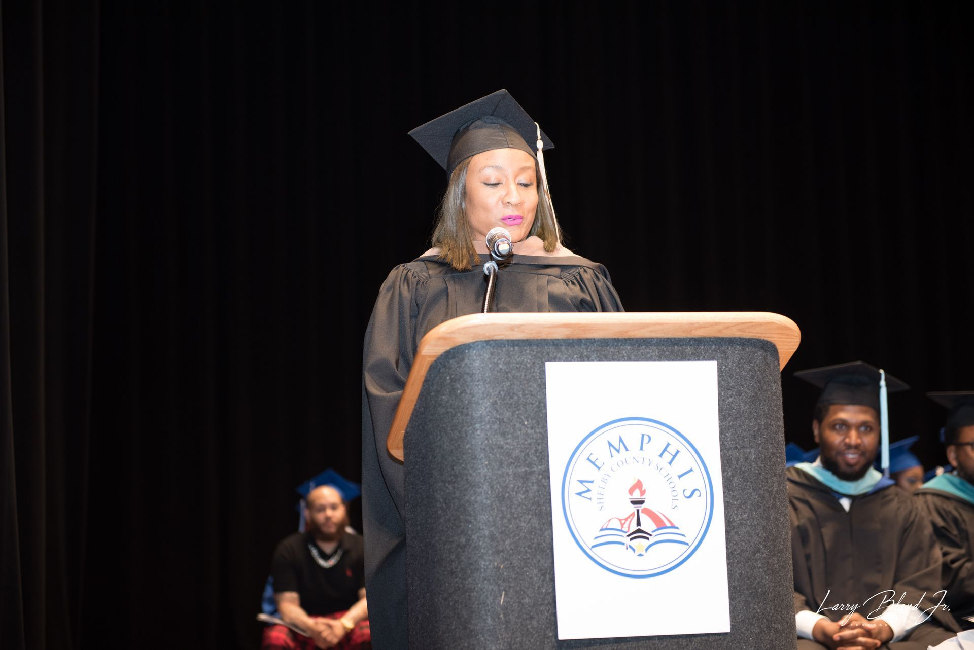 Person in graduation attire speaking at a podium with a school logo, other graduates in background.