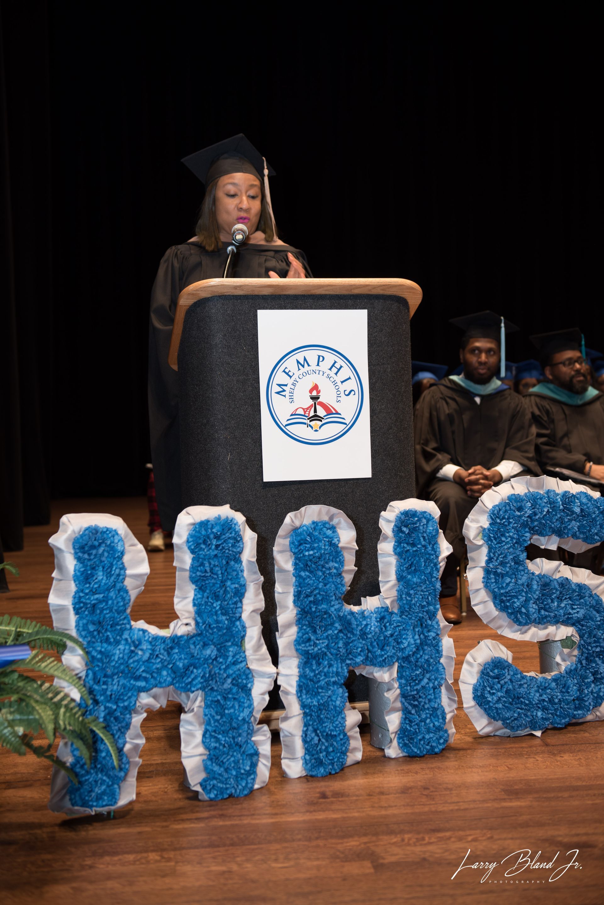 A graduate in cap and gown speaks from a podium with a school logo. 