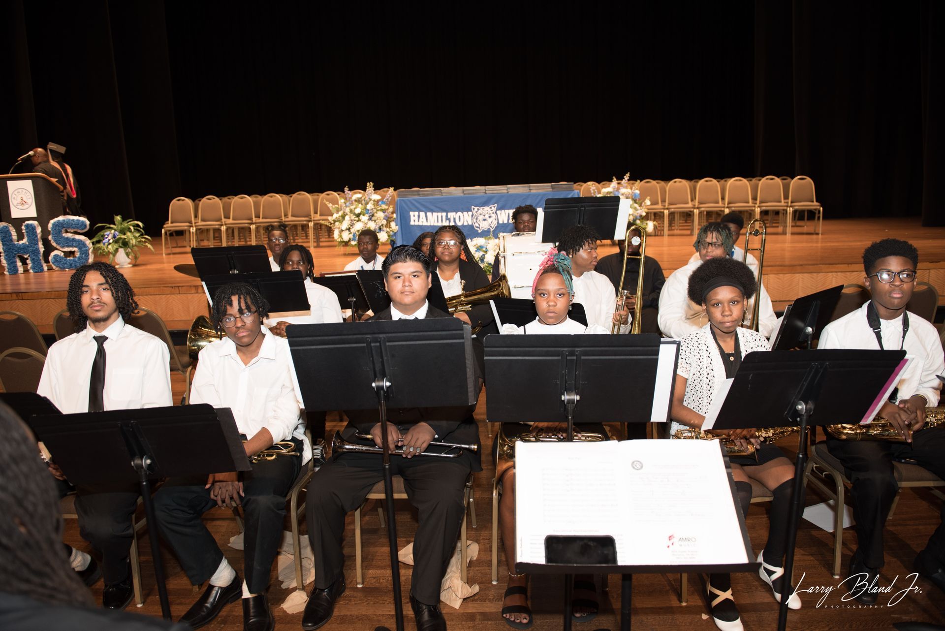 High school band performing on stage. Musicians in formal attire seated, playing instruments, in an auditorium.