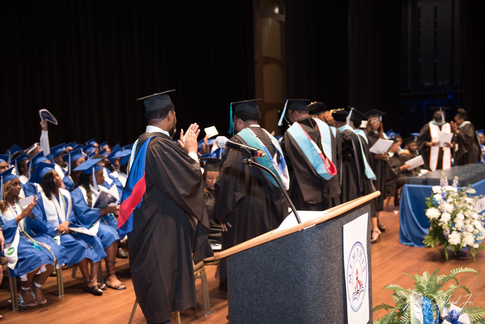 Graduation ceremony with graduates in caps and gowns. Blue and white colors are present.