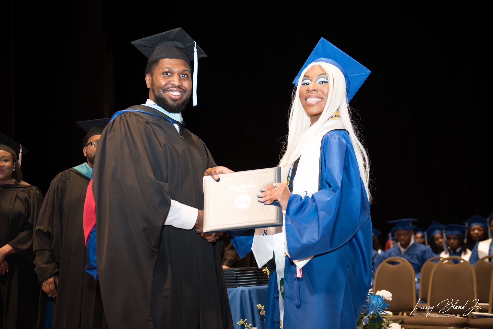 A graduate in blue robe receives a diploma from a person in a black gown at a ceremony.