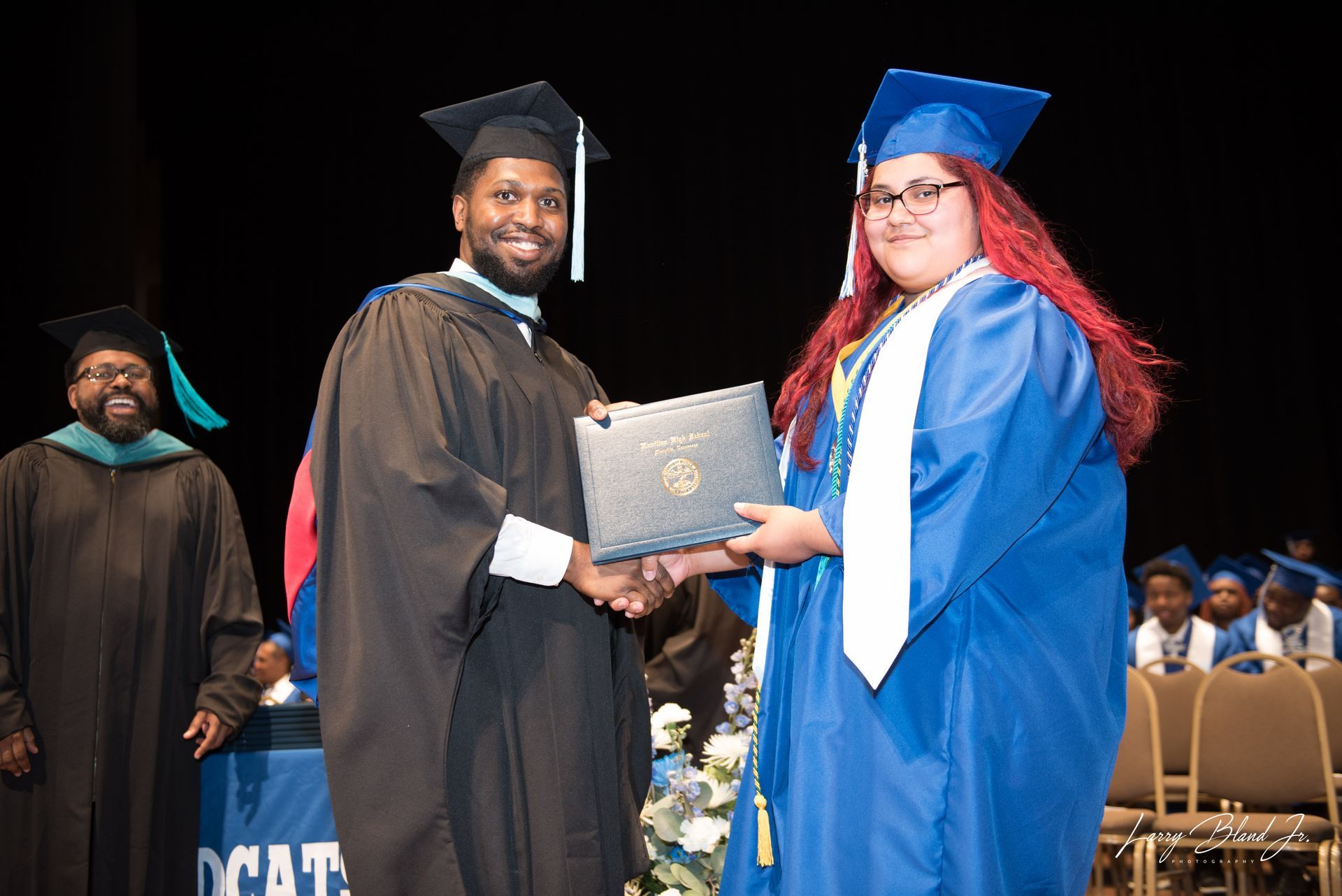 Man in black gown hands diploma to student in blue gown onstage.