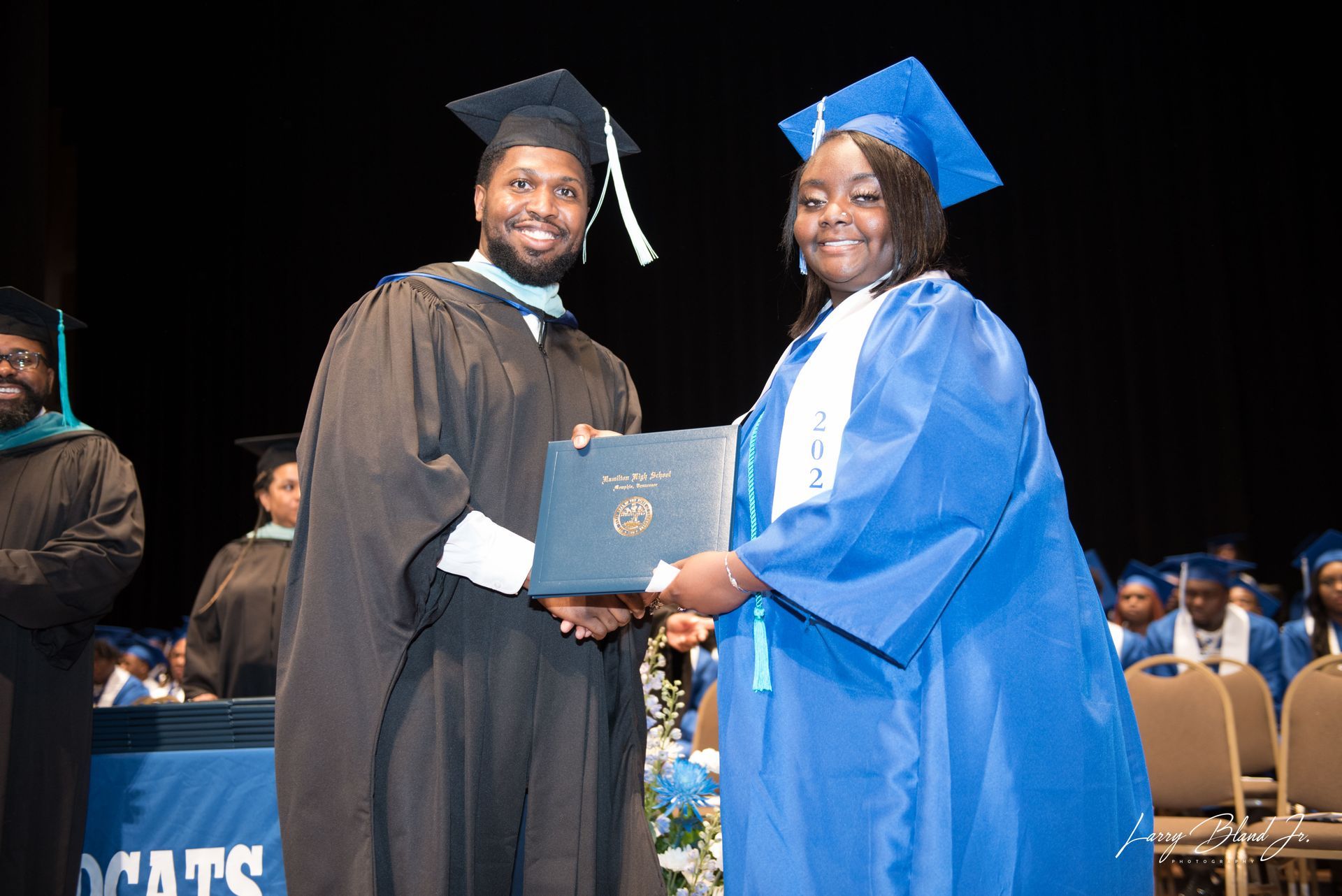 Graduation ceremony: Man in black robe hands diploma to woman in blue robe.