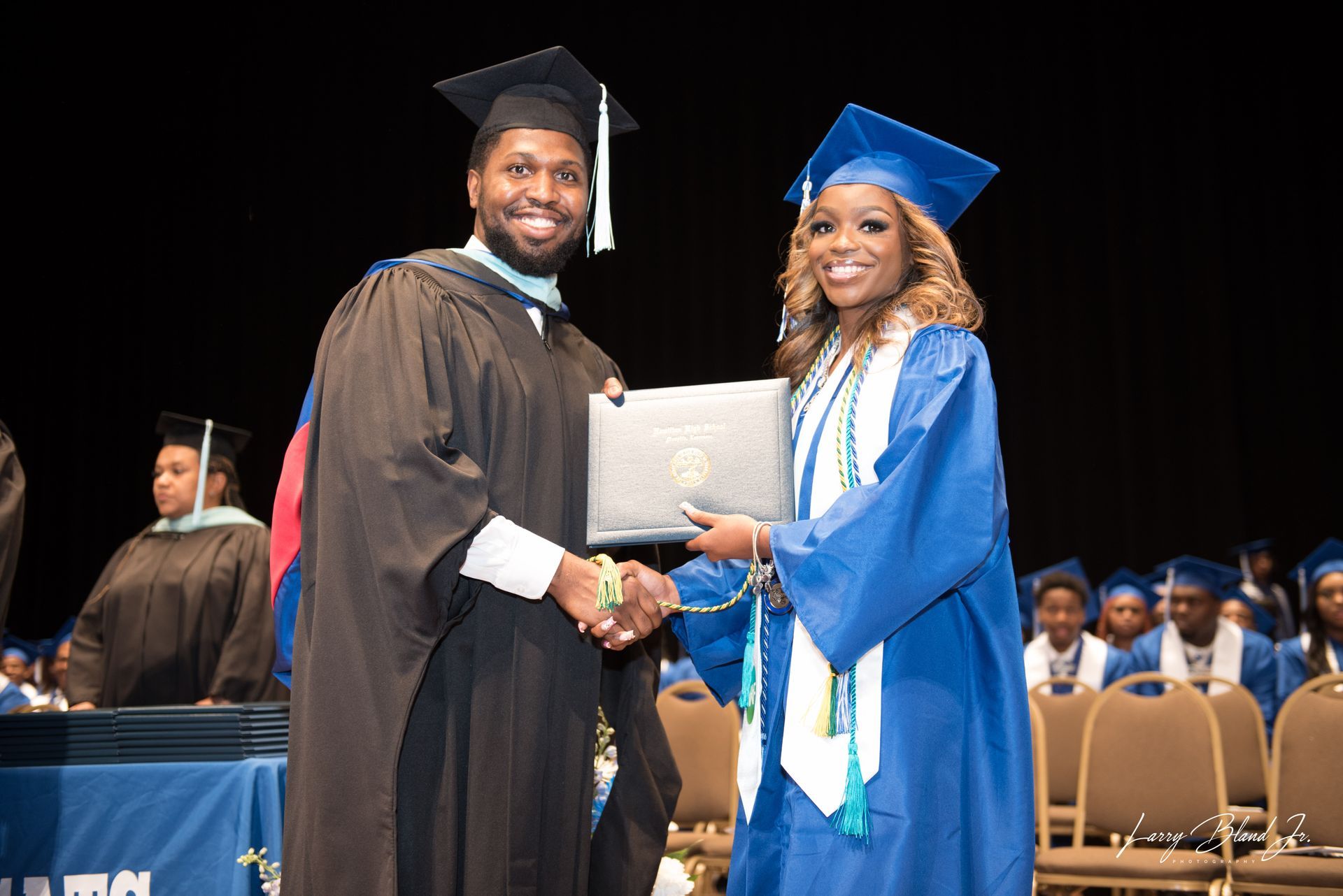 Person in black robe handing diploma to person in blue gown; graduation ceremony.